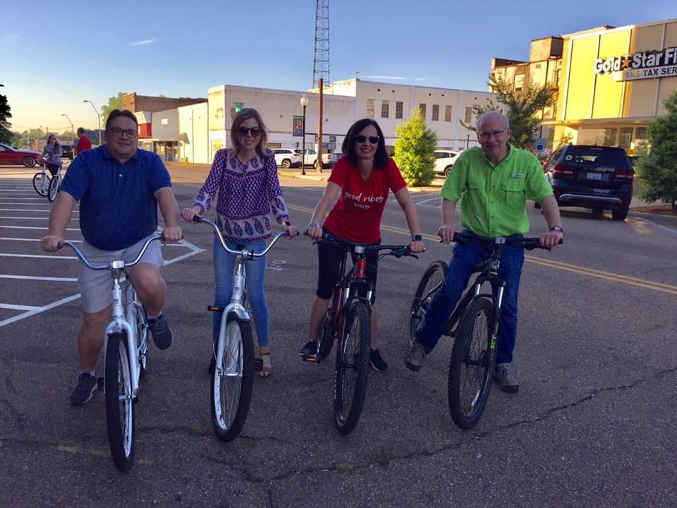 Four people ride bicycles on a street in a small town. Buildings and a tower are in the background.