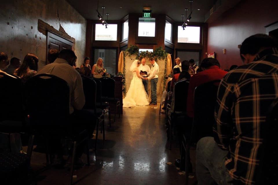 Couple getting married inside, in front of an archway; guests seated on either side, dark interior, event.