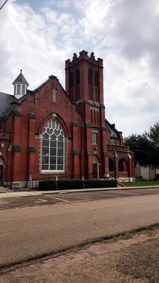 Brick church with a tall tower and large stained-glass window under a cloudy sky.