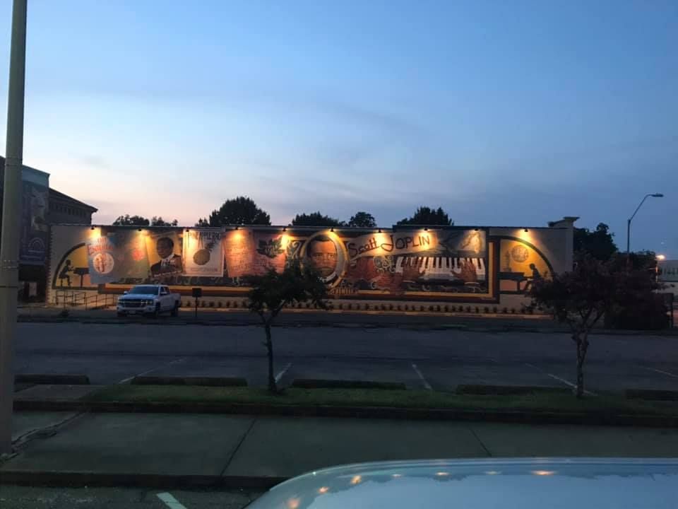 Mural on building featuring musical instruments and portraits, lit at dusk. Parked vehicles and street lights in front.