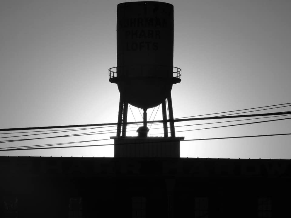 Silhouette of a water tower atop a building, against a bright sky, with power lines in the foreground.