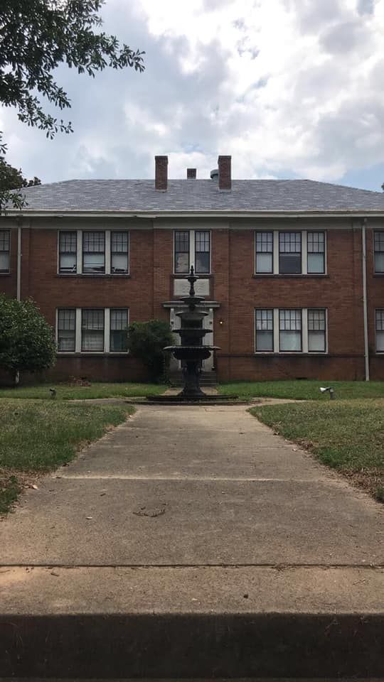 Brick apartment building with a tiered fountain in front; gray roof and cloudy sky.