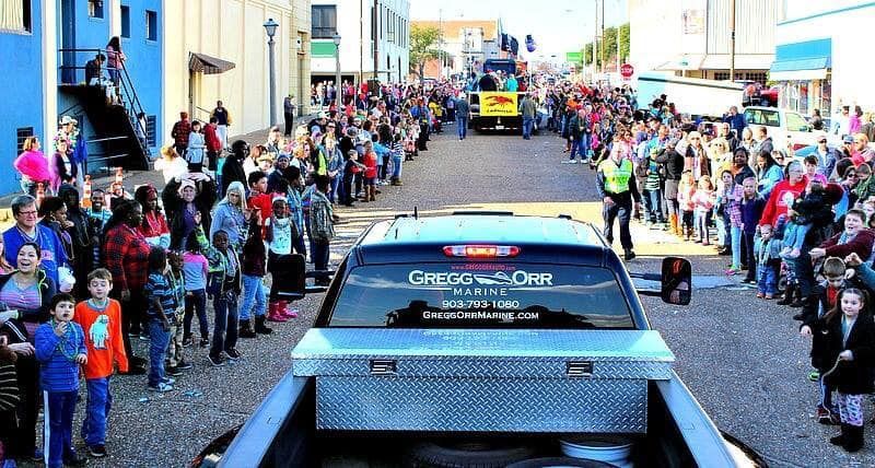 Parade with crowds lining the street; a truck with a toolbox leads. Buildings and blue sky in background.