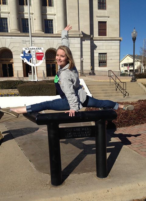 Woman doing splits on a black bench outside a building, arm raised, wearing jeans and a gray hoodie.