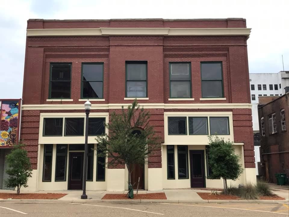 Two-story brick building with large windows and tan trim. Small trees line the sidewalk.