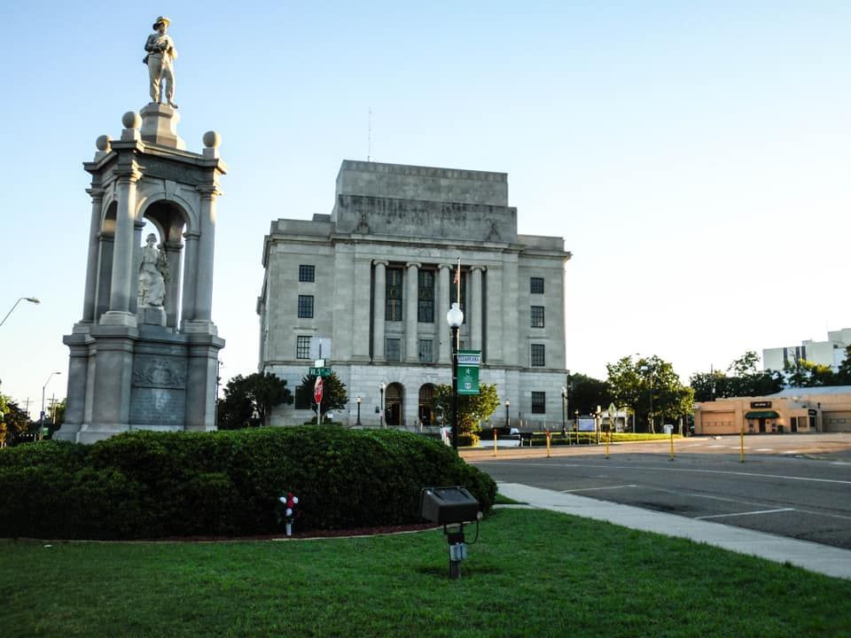Statue in front of a stone courthouse building on a green lawn with a tree and a road.