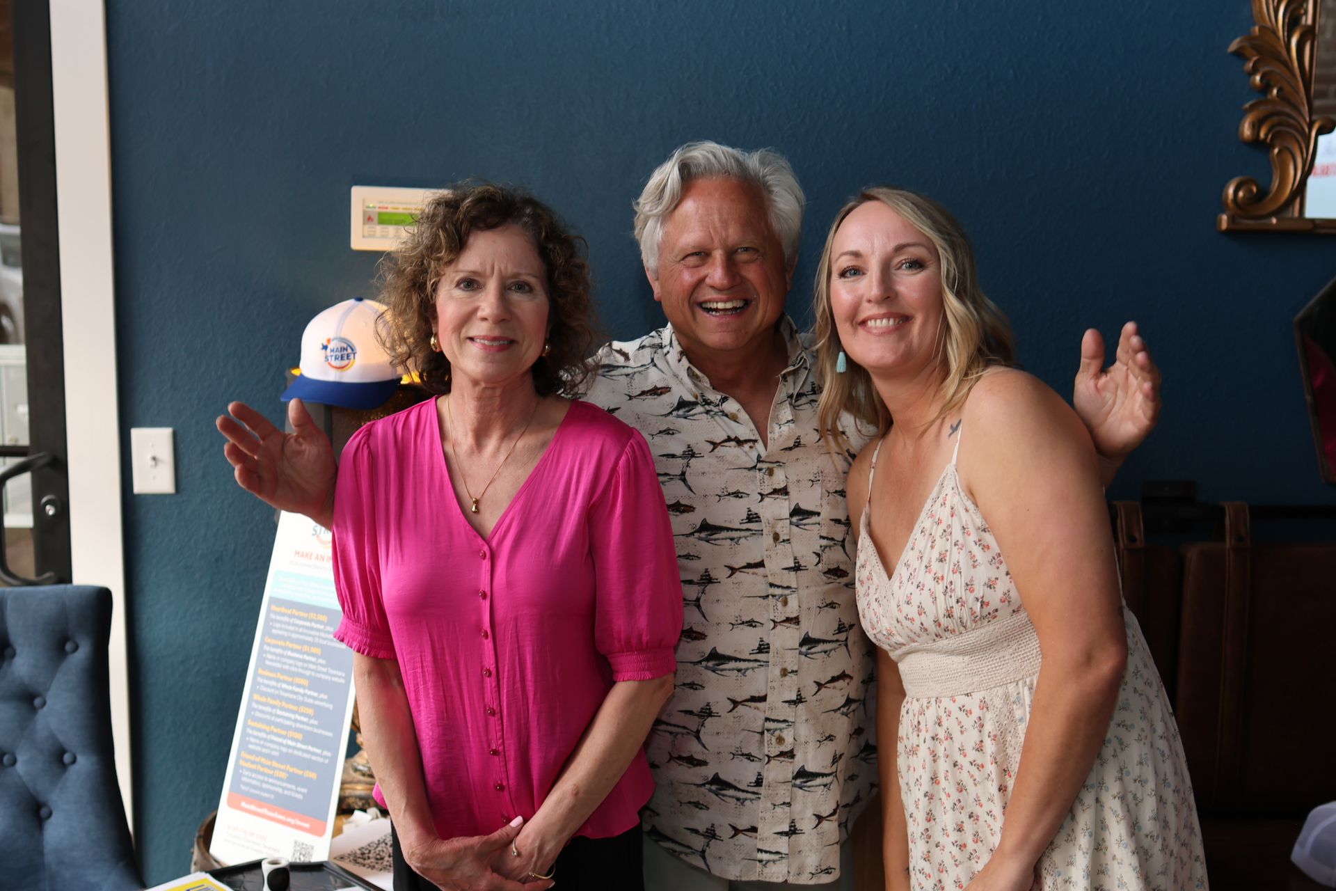 Three smiling people posing indoors, with one woman waving at the camera.