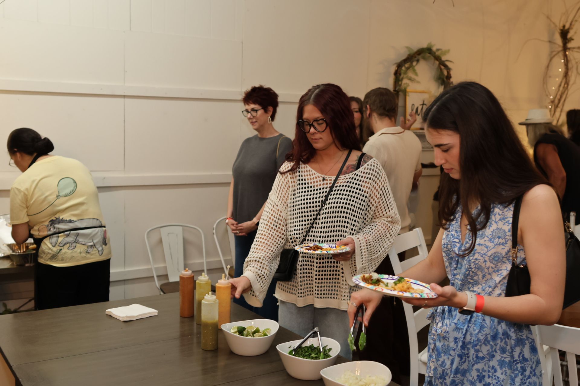 People serving food at a buffet table indoors, with bowls, plates, and condiments.