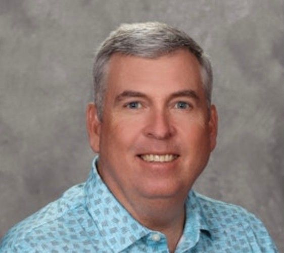 Studio headshot of a smiling man in a light blue patterned shirt against a gray backdrop