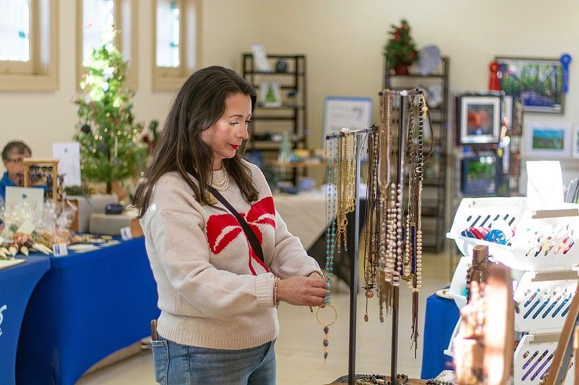 Woman browsing necklaces at an indoor craft fair. She wears a sweater with a bow design.