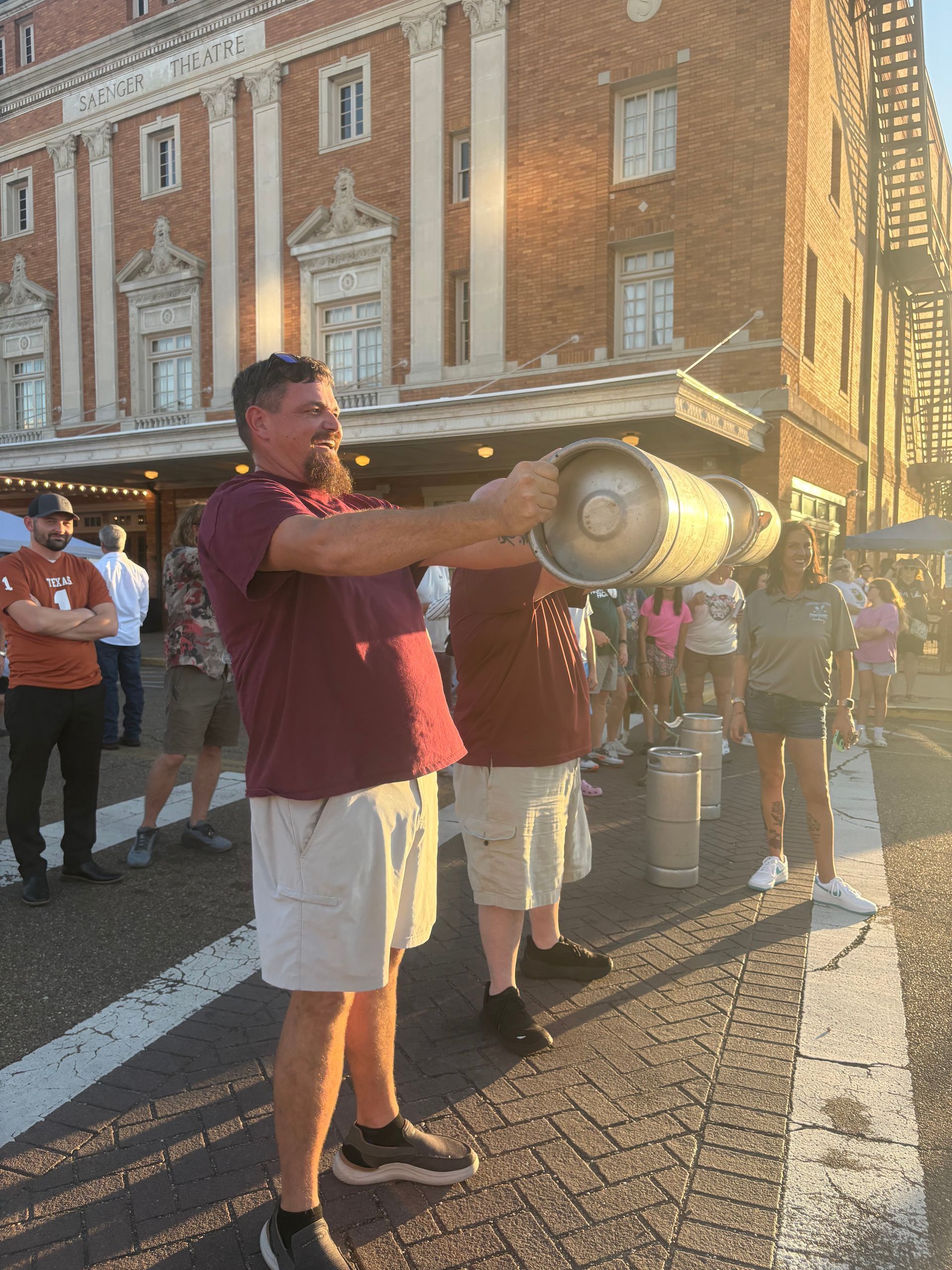 Man holding a large metal keg in front of a building with others nearby; sunny outdoors.