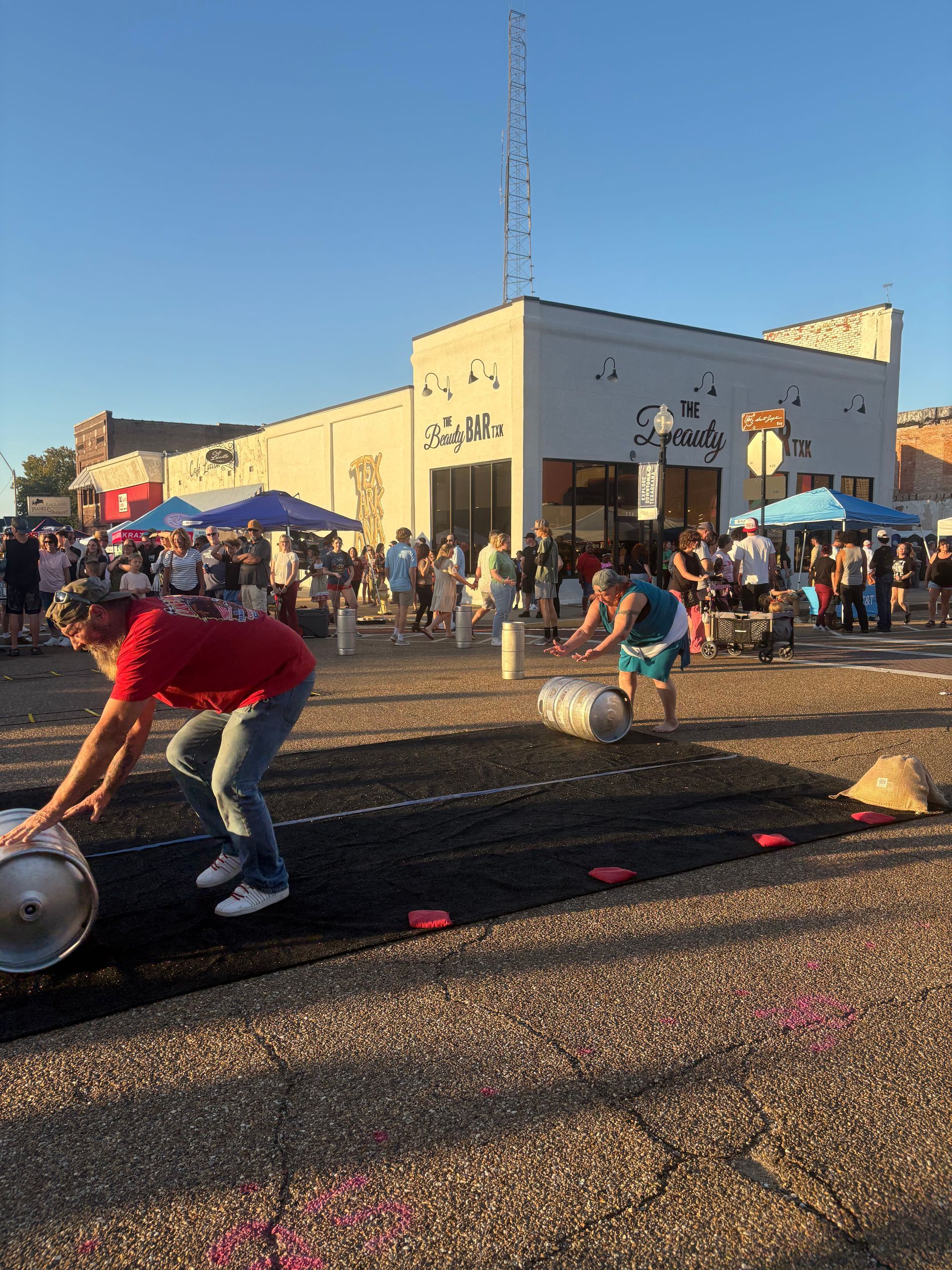 People rolling barrels on a black track at an outdoor event near a white building.