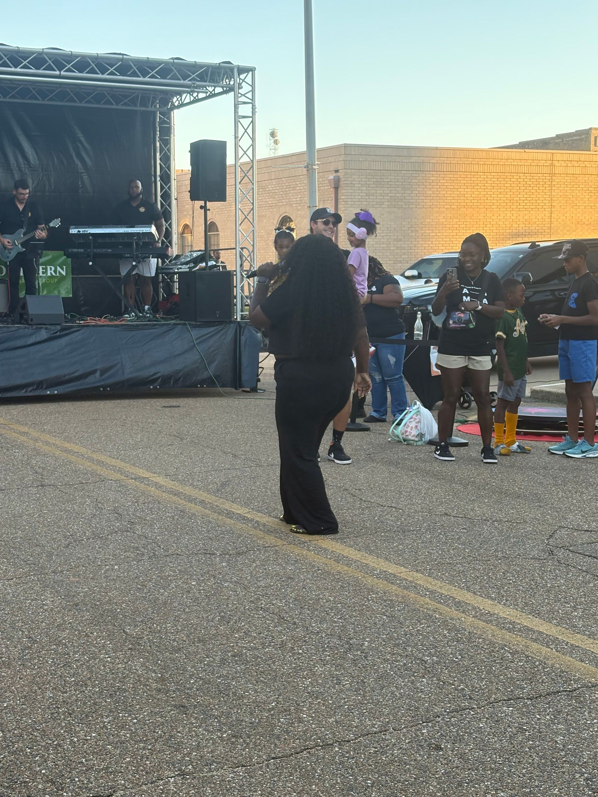 Person dancing in front of a band on stage at an outdoor event. People watch nearby.