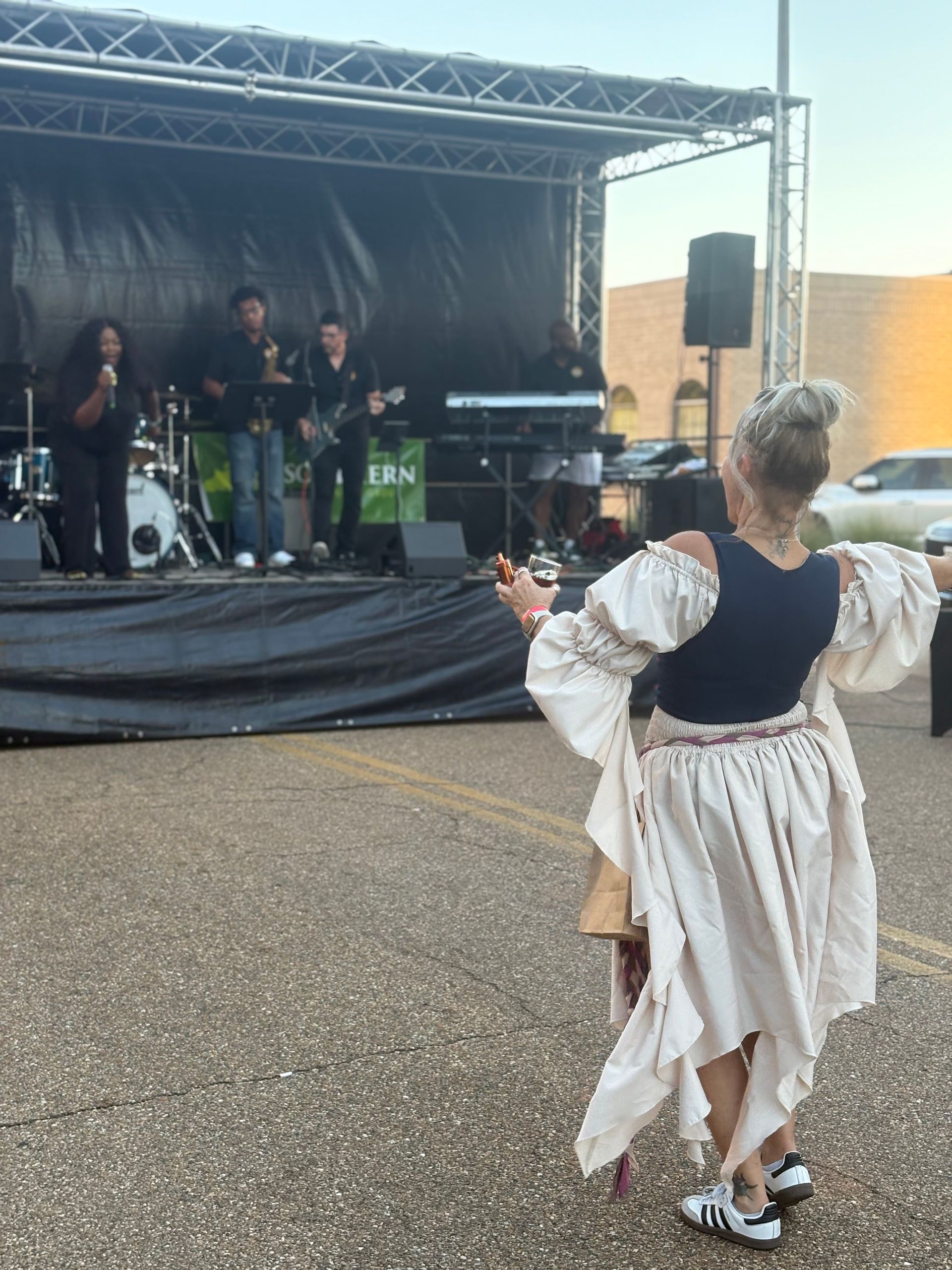 Woman dancing in front of a band on stage, outdoors. The band consists of multiple musicians.