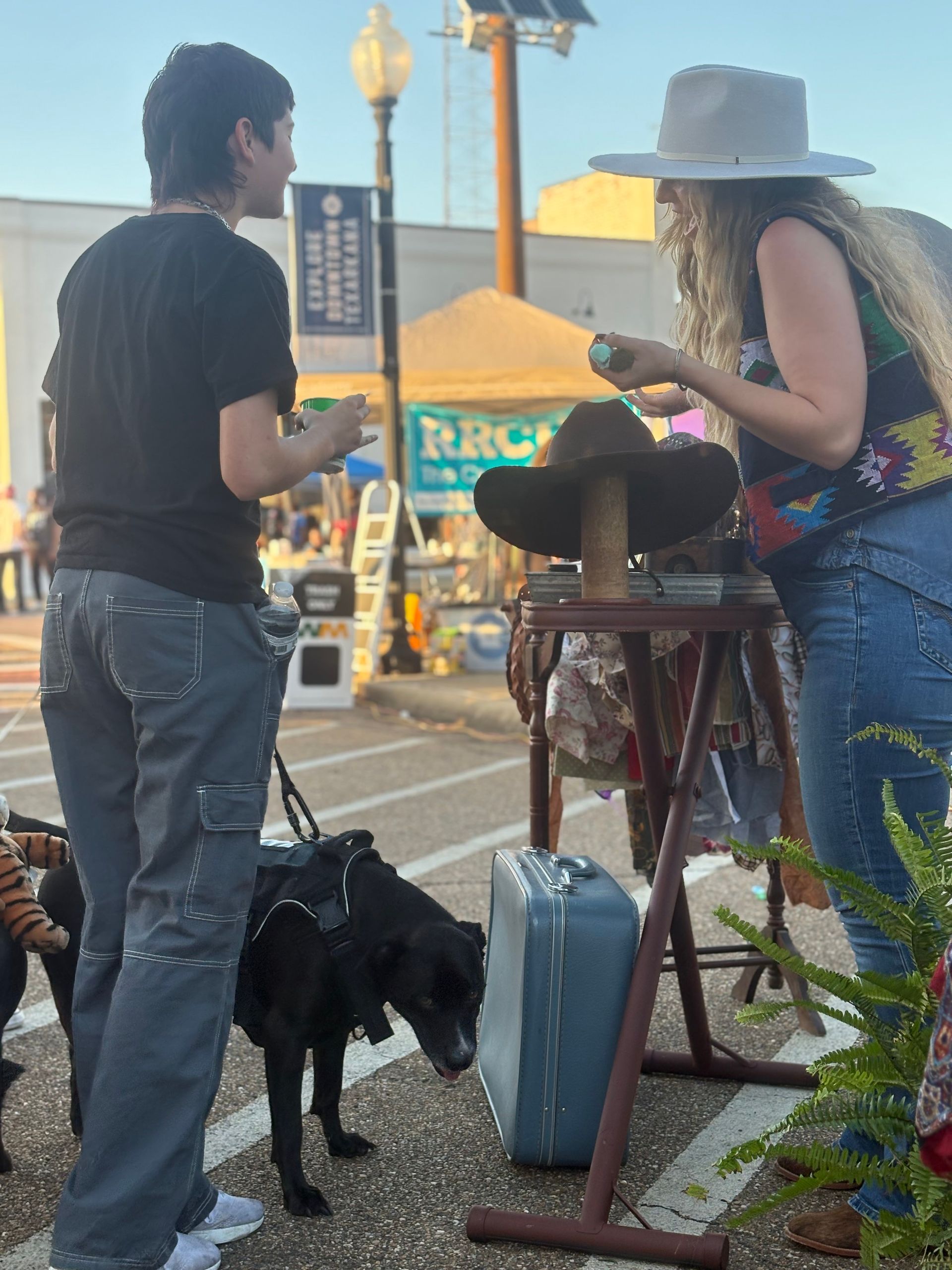 Person with dog shopping at outdoor vendor. Woman examines hats, while a black dog sniffs a vintage suitcase.