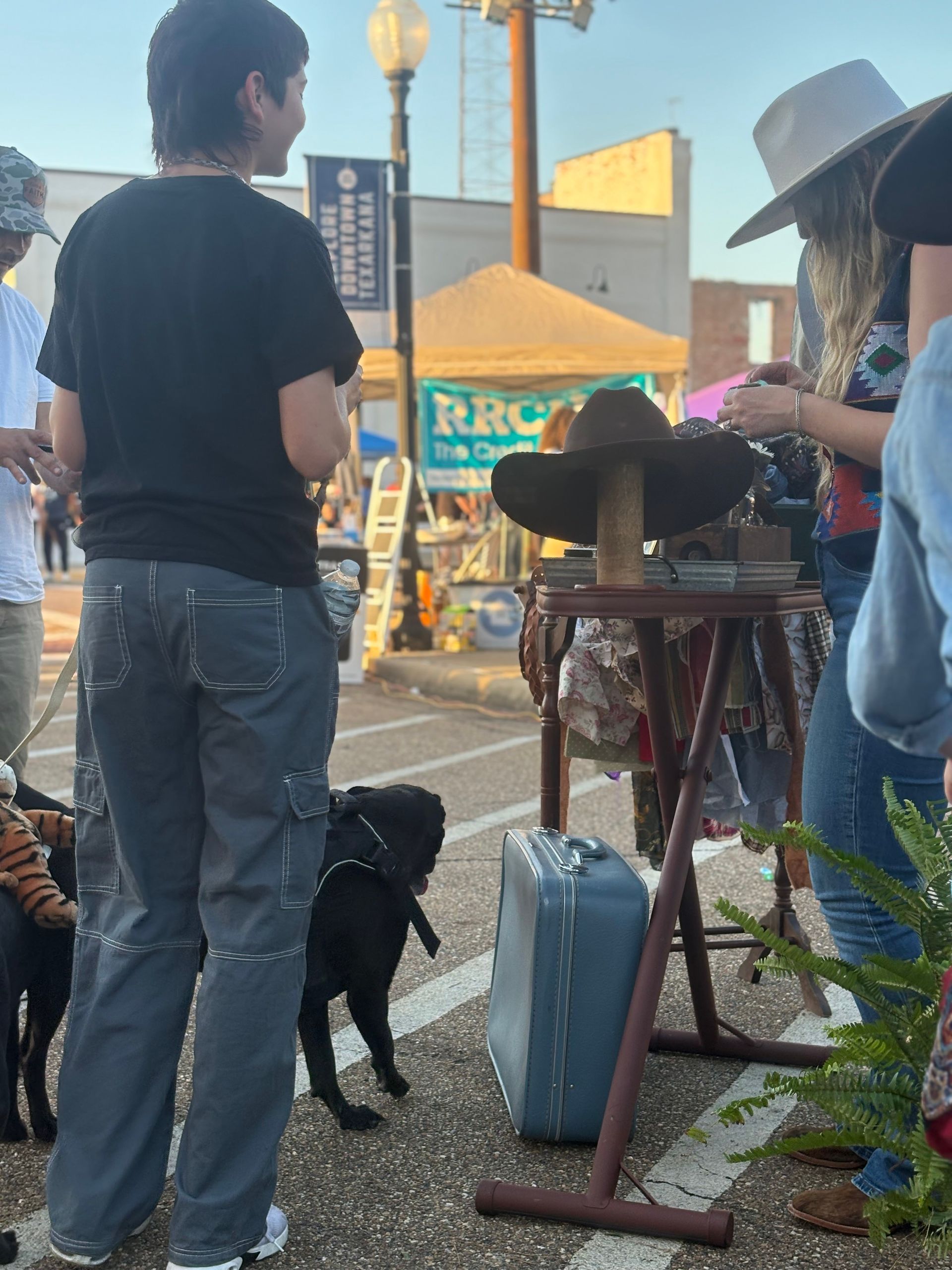 People browsing an outdoor market. A dog walks by a display of hats and clothing. A blue suitcase is nearby.
