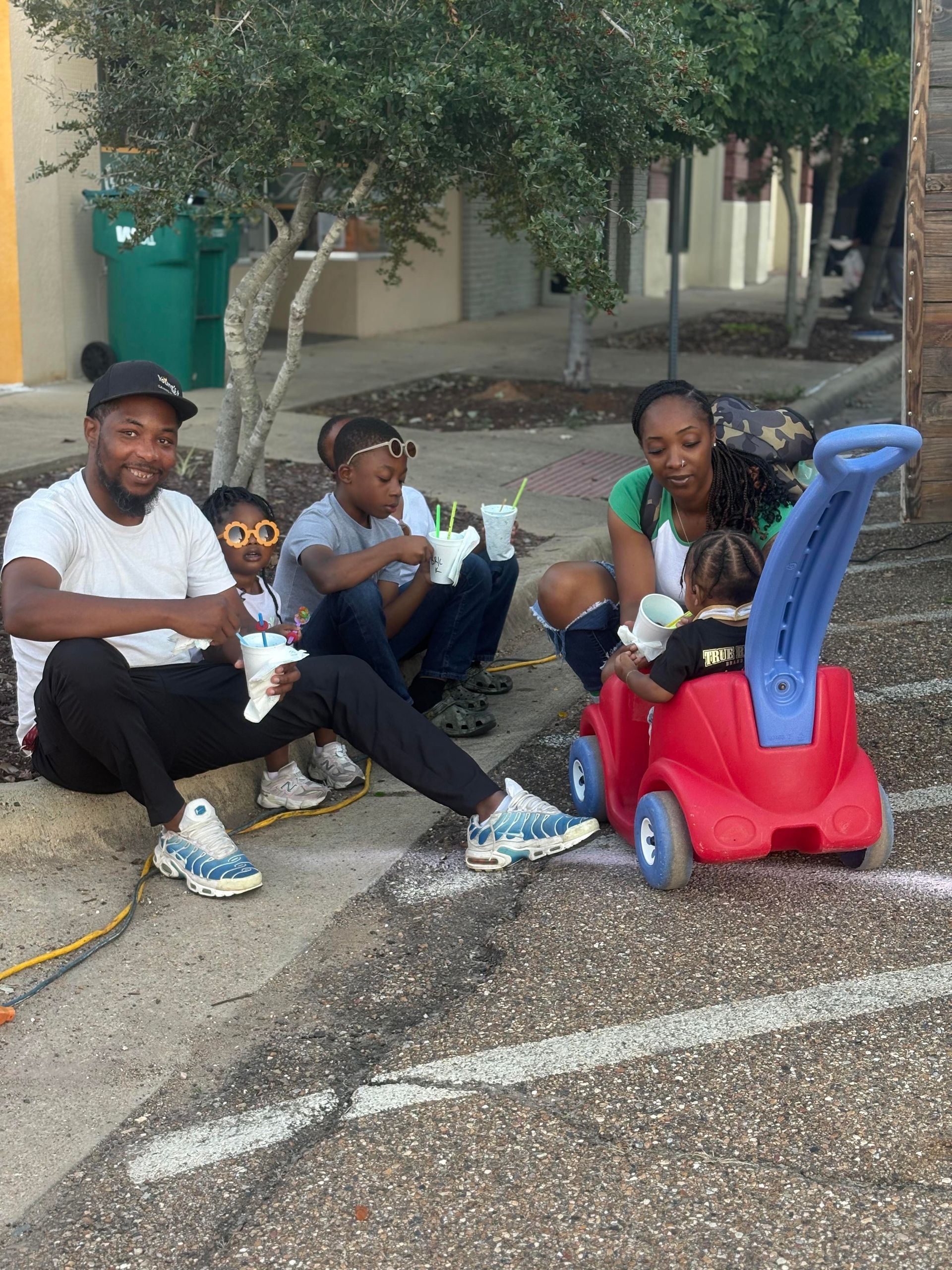 Family sitting on curb with drinks, toddler in toy car. Buildings and trees in the background.