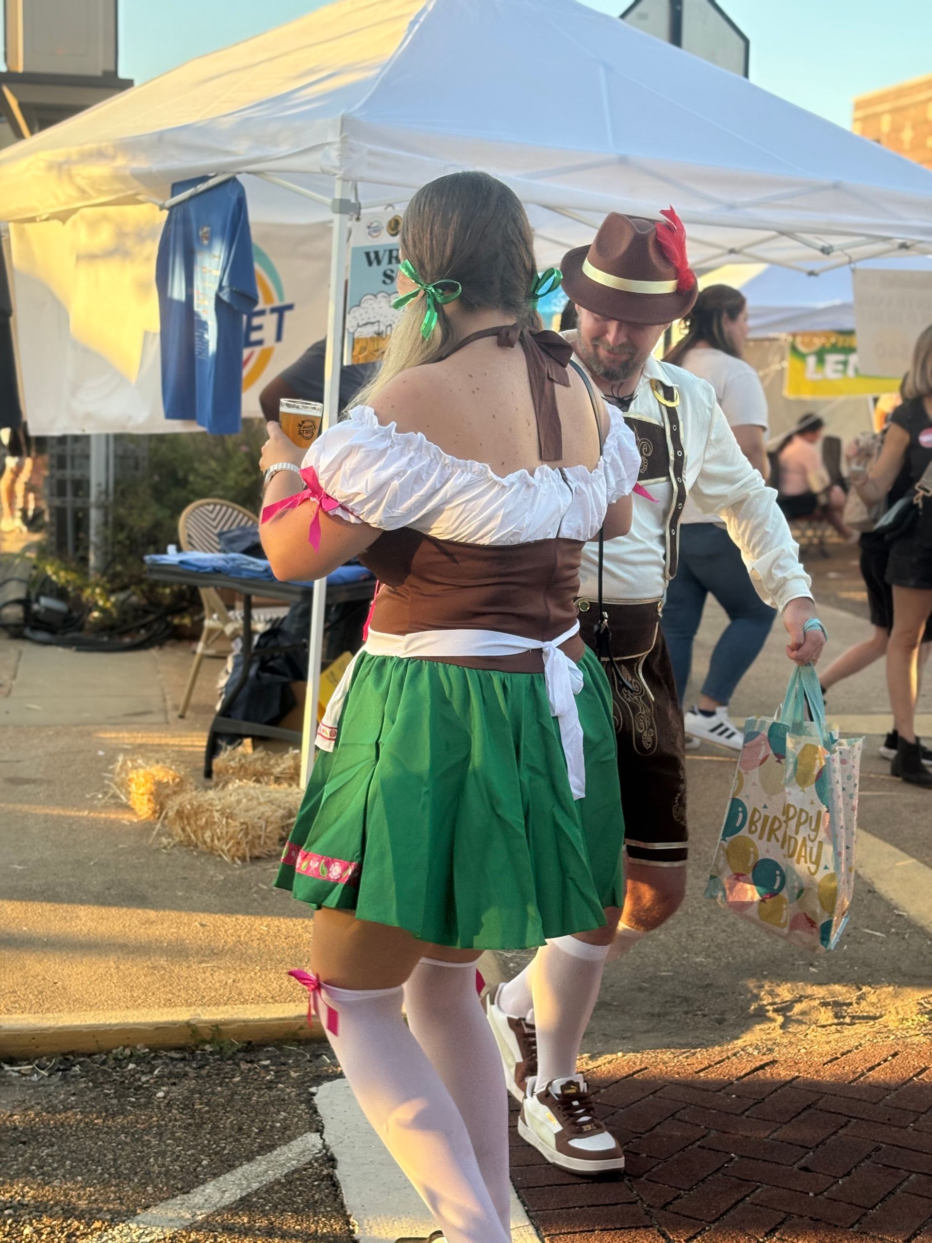 People in traditional German outfits, dancing at an outdoor event with a white tent in the background.