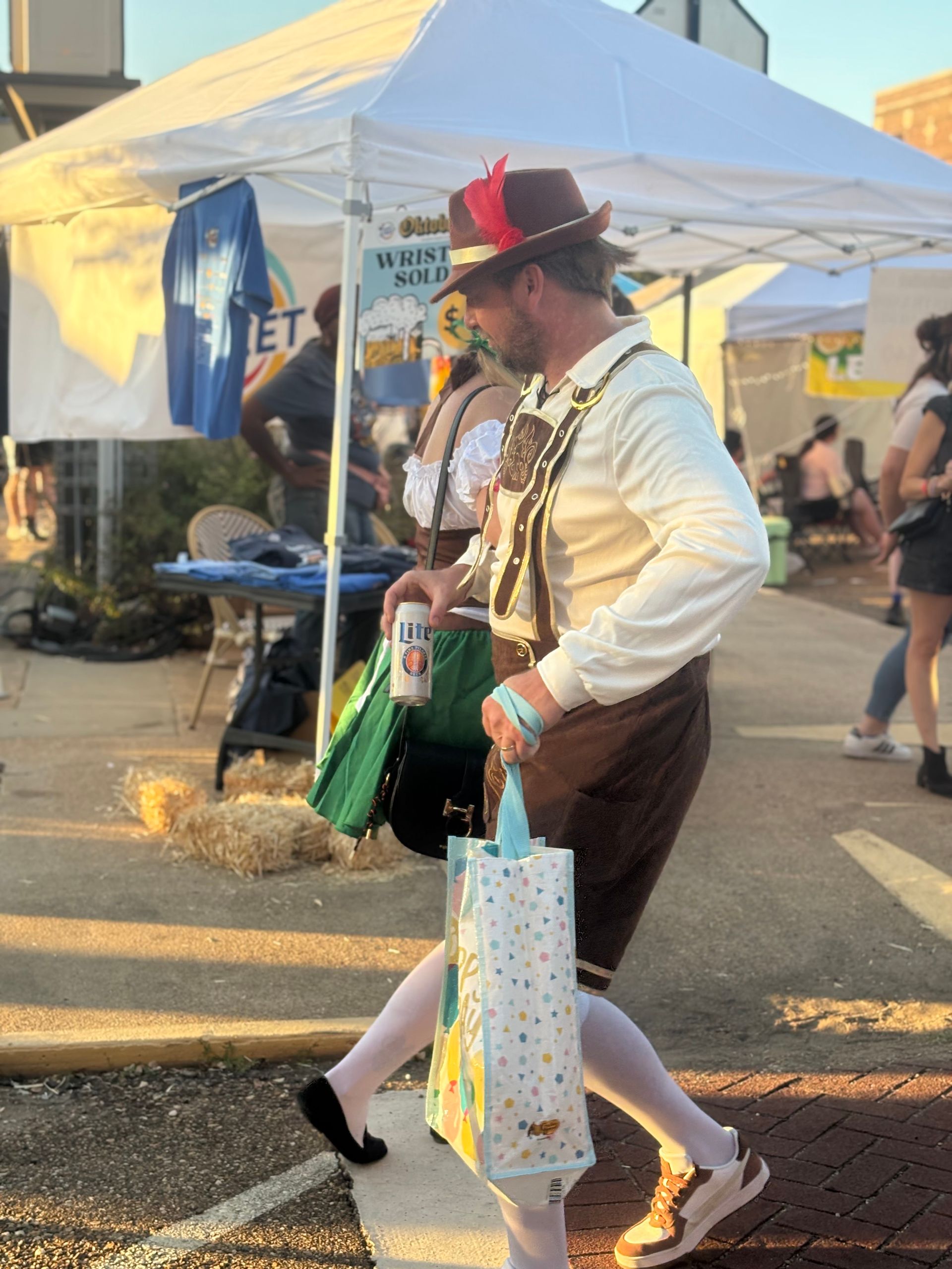 Man in lederhosen walking with a beer and gift bag at an outdoor event with tents.