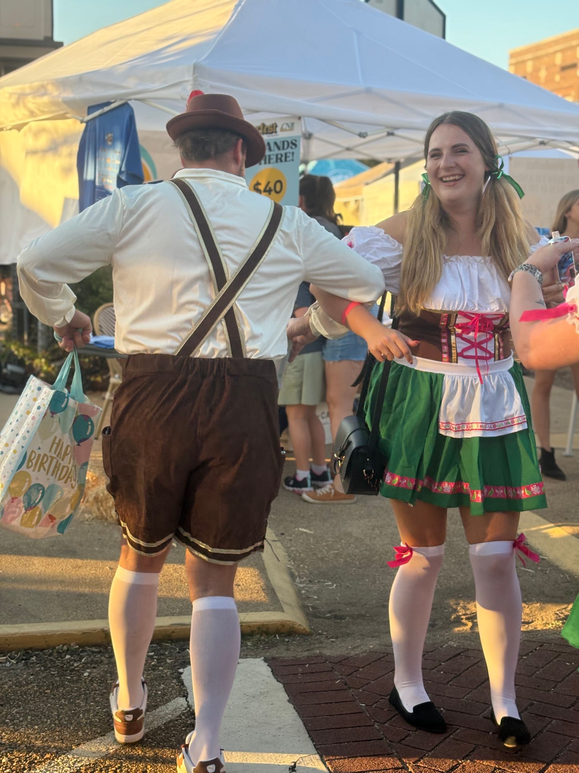 Two people in Oktoberfest attire dancing outdoors near a white tent.