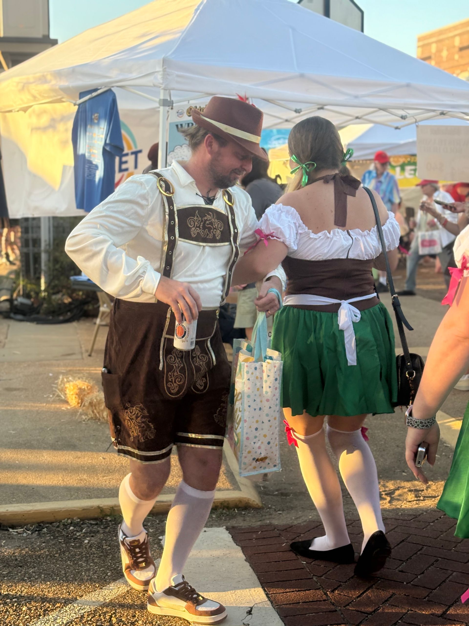 People in traditional German attire at an outdoor event, man with beer, woman with gift bag.