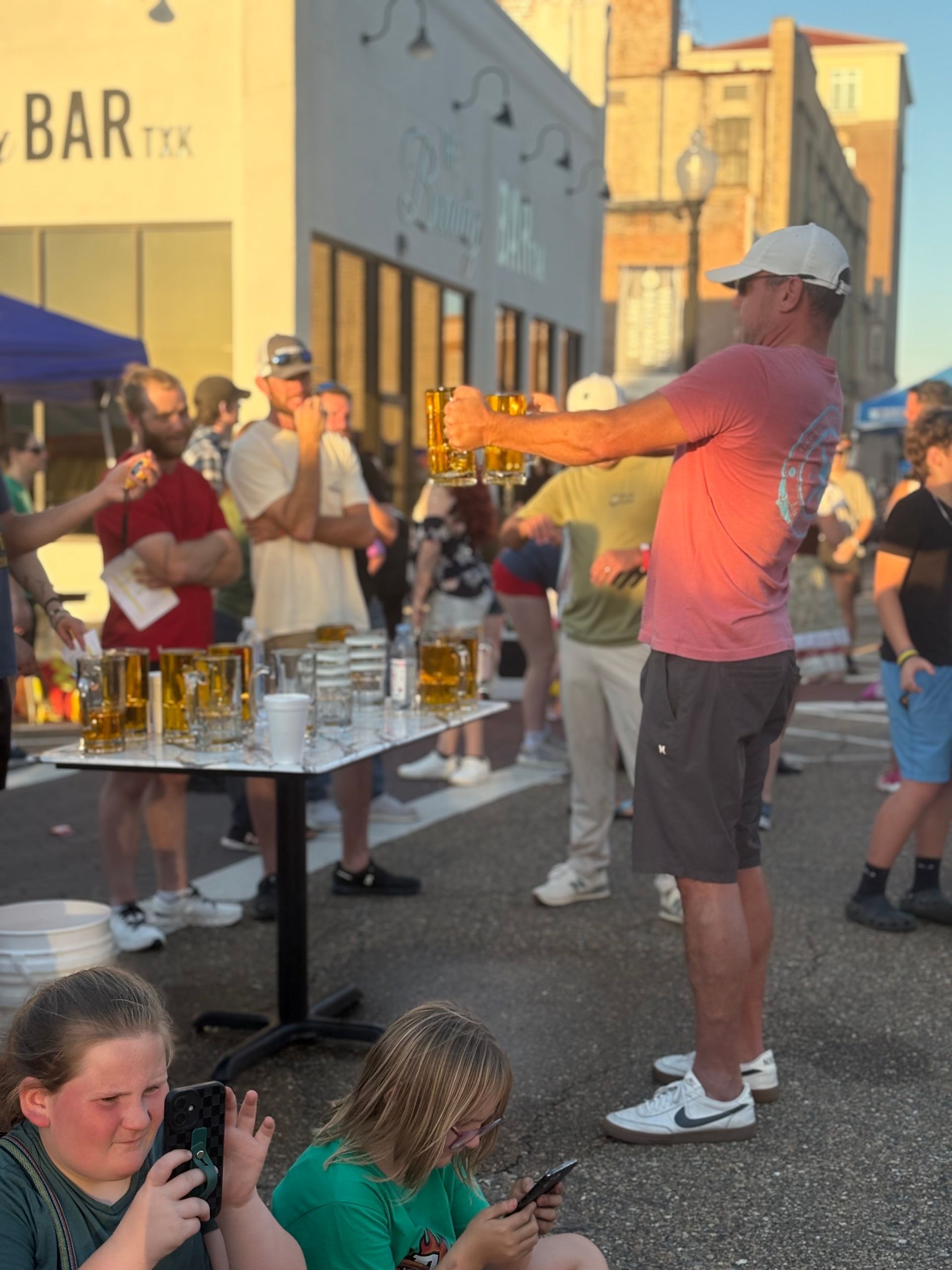 Man holding many full beer glasses at outdoor event. People watch.