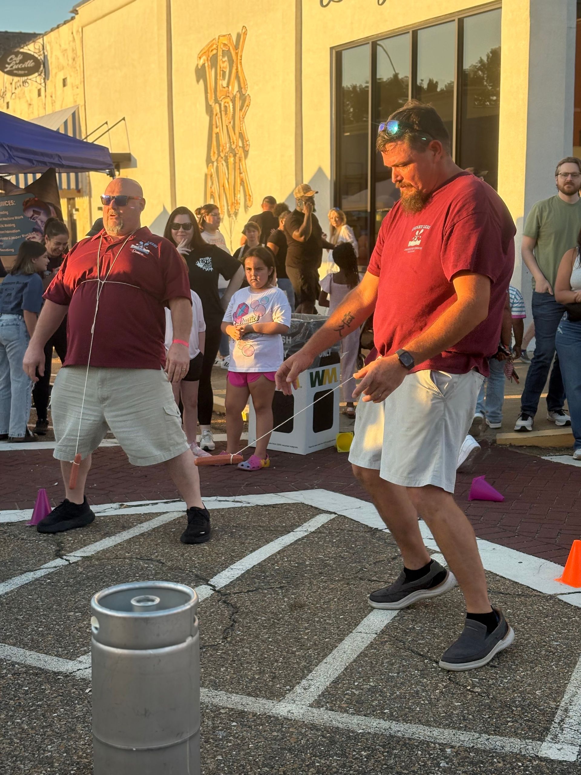 Two men playing a game outdoors. They wear maroon shirts, khaki shorts, and face each other near a metal cylinder.