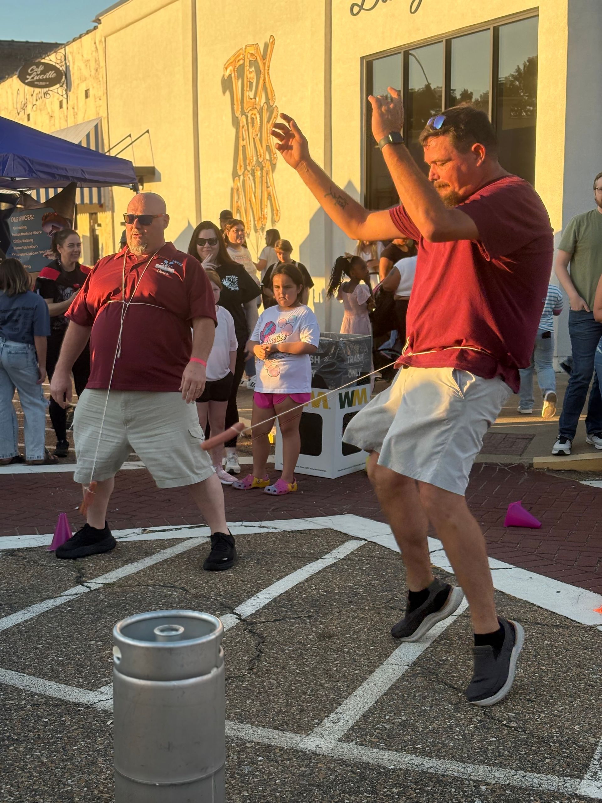 Two men playing a game outdoors. One jumps, arms raised. Both wear maroon shirts and khaki shorts. A crowd watches.