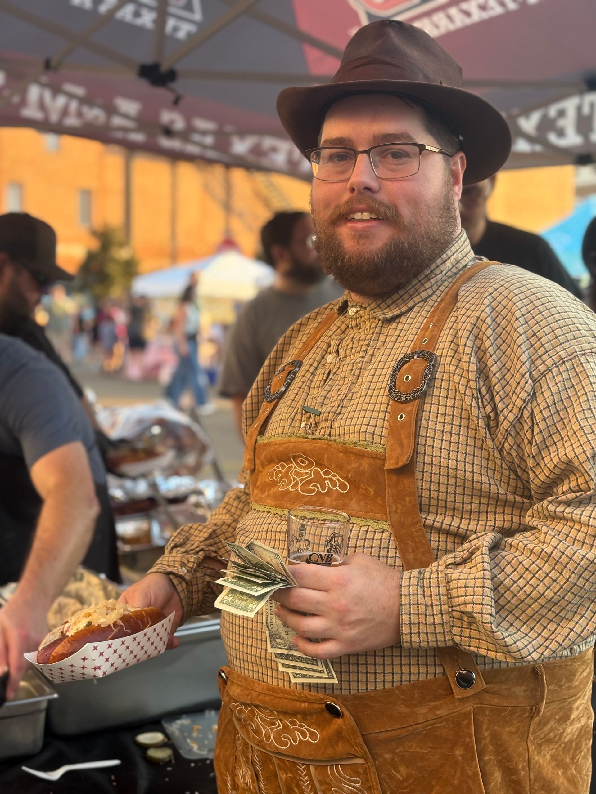 Man in brown hat and apron holding food and money at a food stall, possibly an outdoor market.