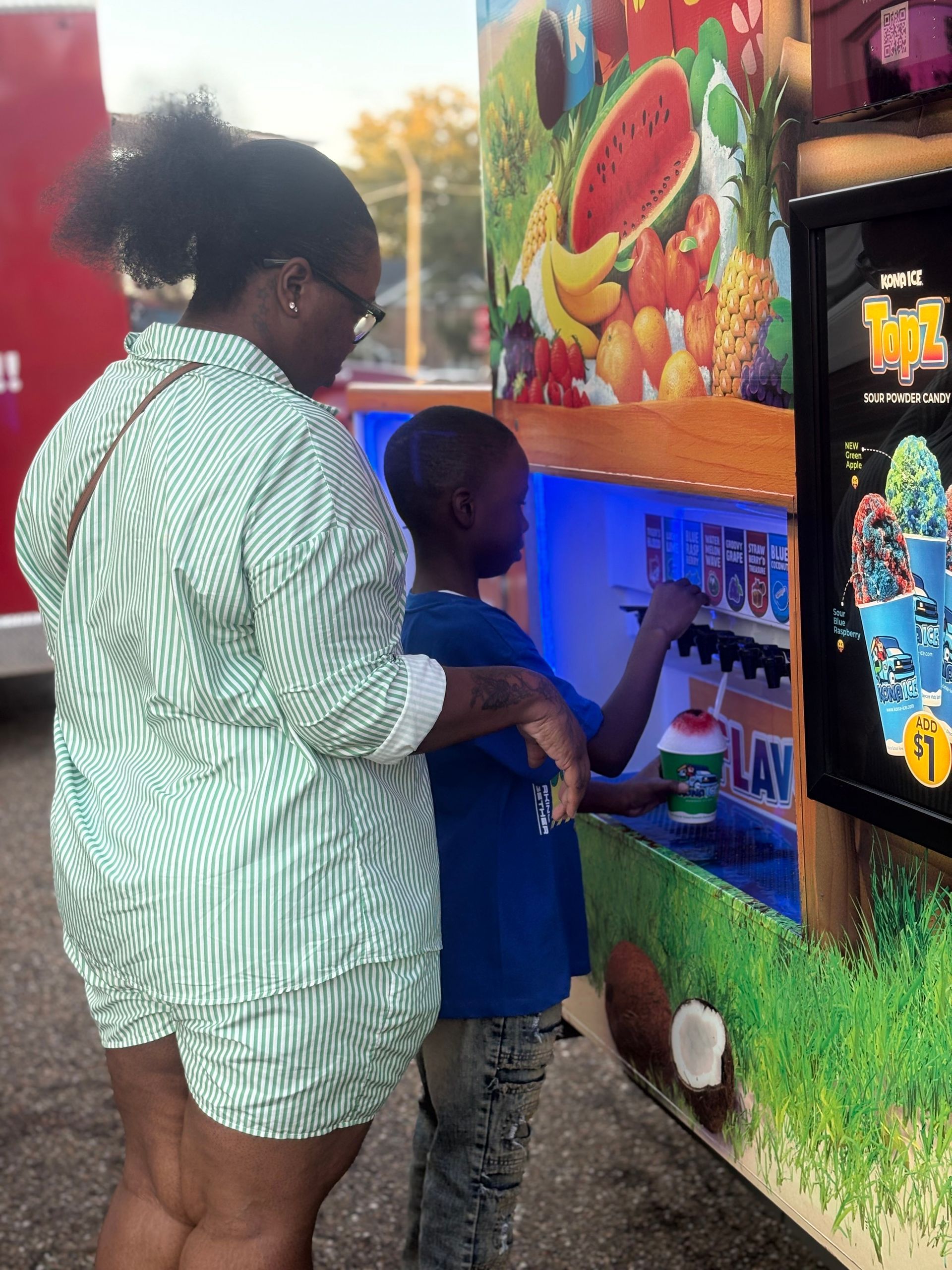 Woman and child at a shaved ice truck. The child dispenses the ice while the woman watches.