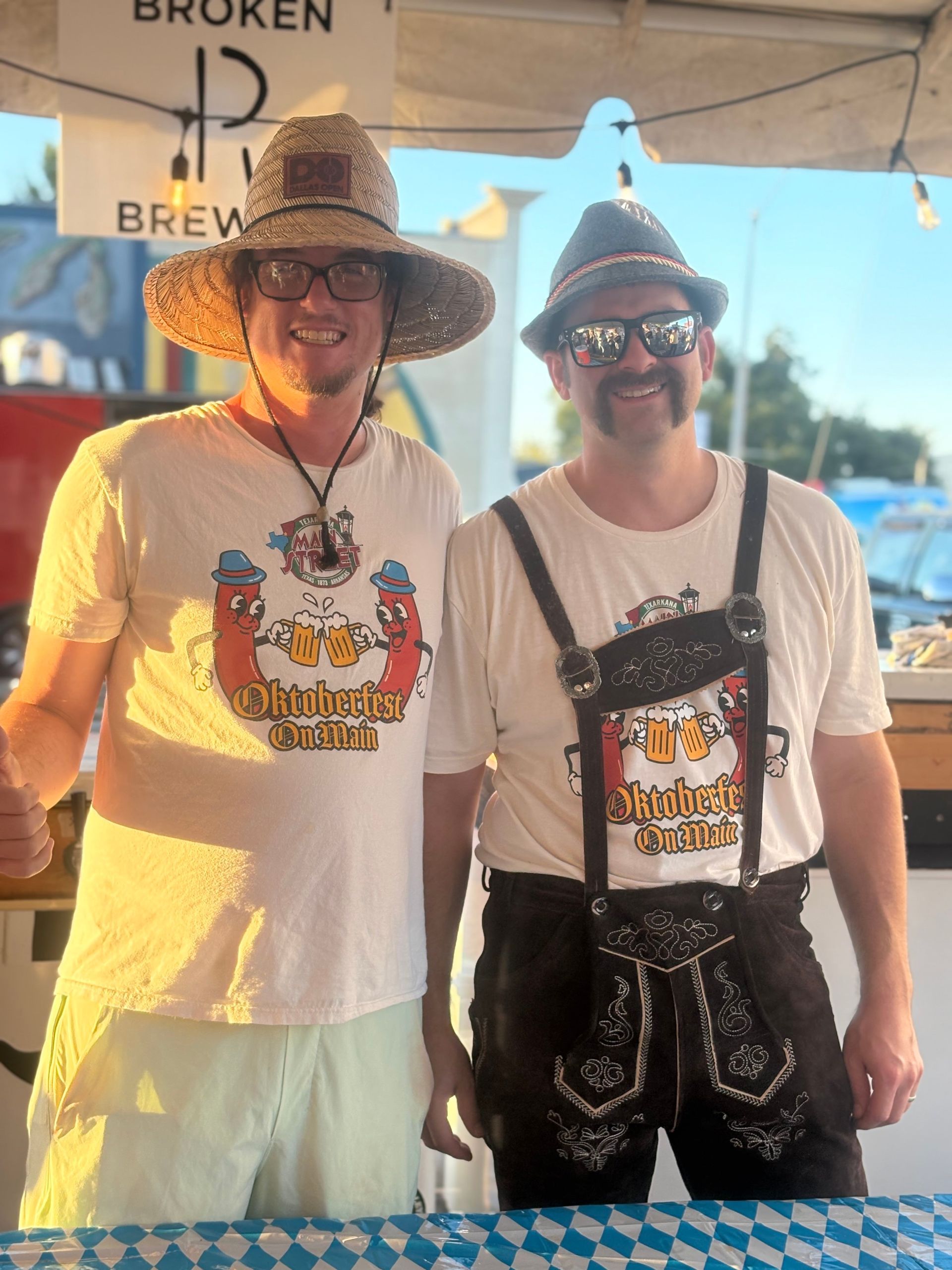 Two men at a brewery stand, one in a straw hat and one in lederhosen.