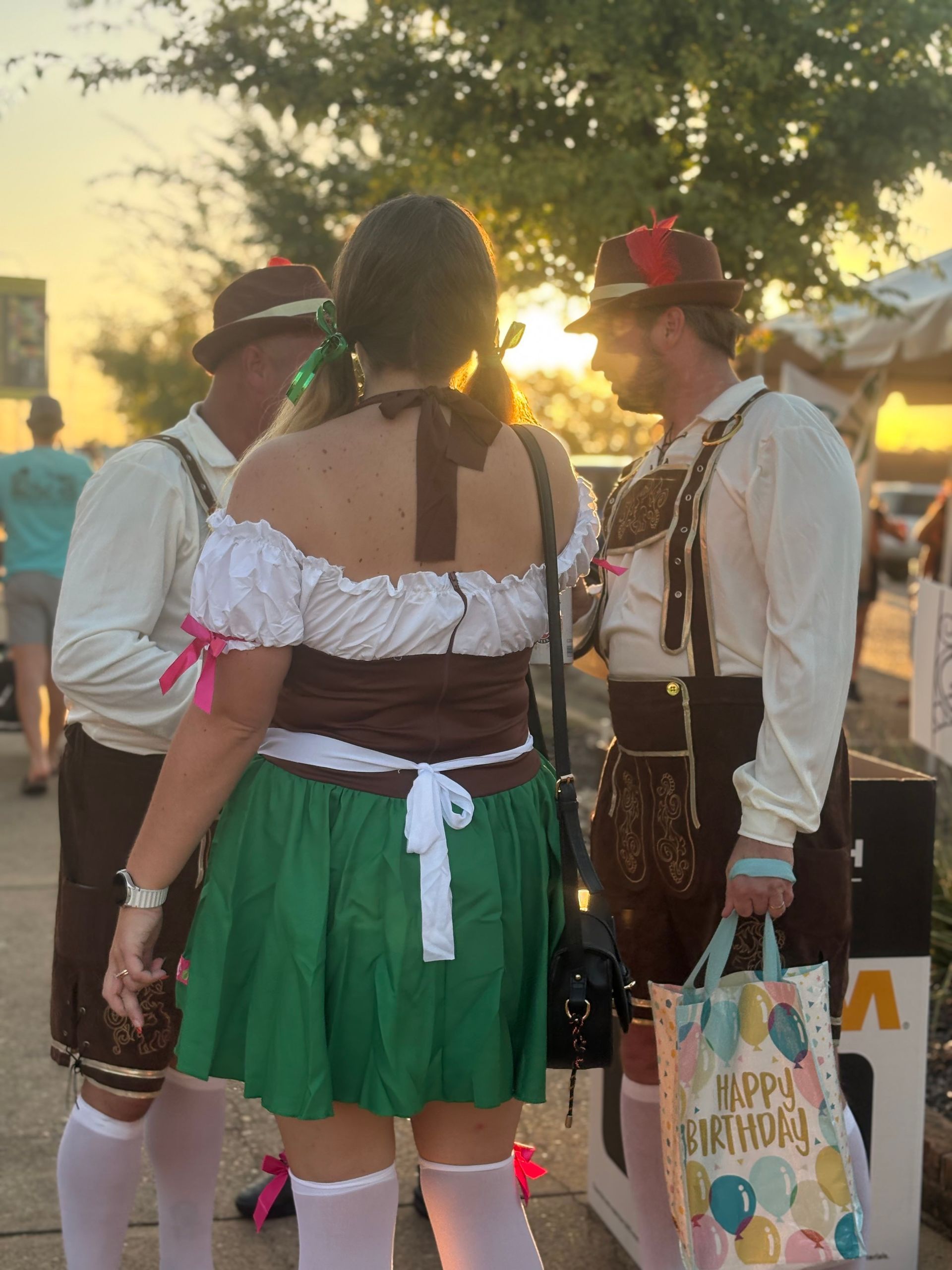 Three people in Bavarian outfits outdoors, possibly at a festival. One holds a 