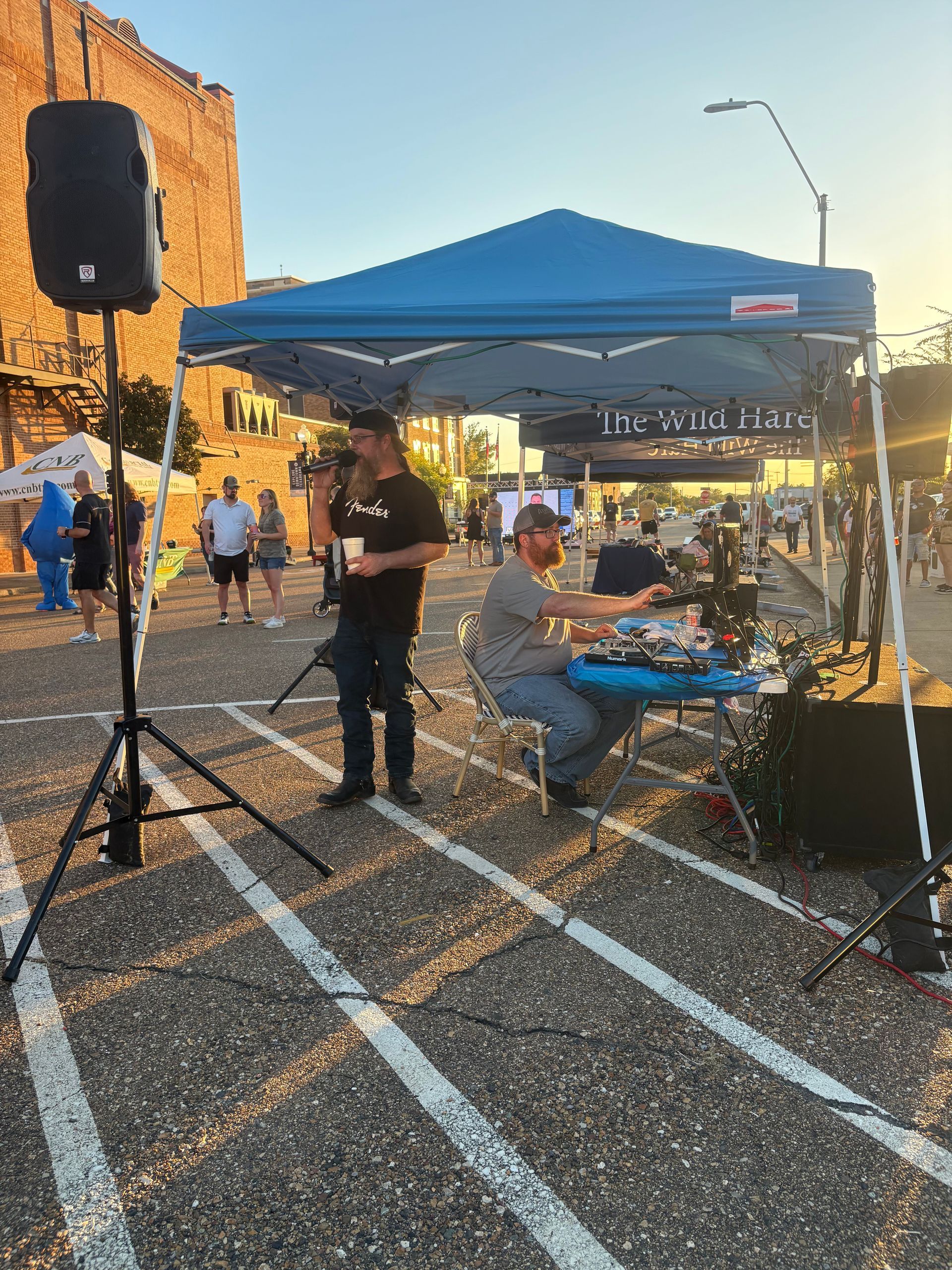 Two men under a blue tent with sound equipment; one speaks into a mic, the other works a soundboard.