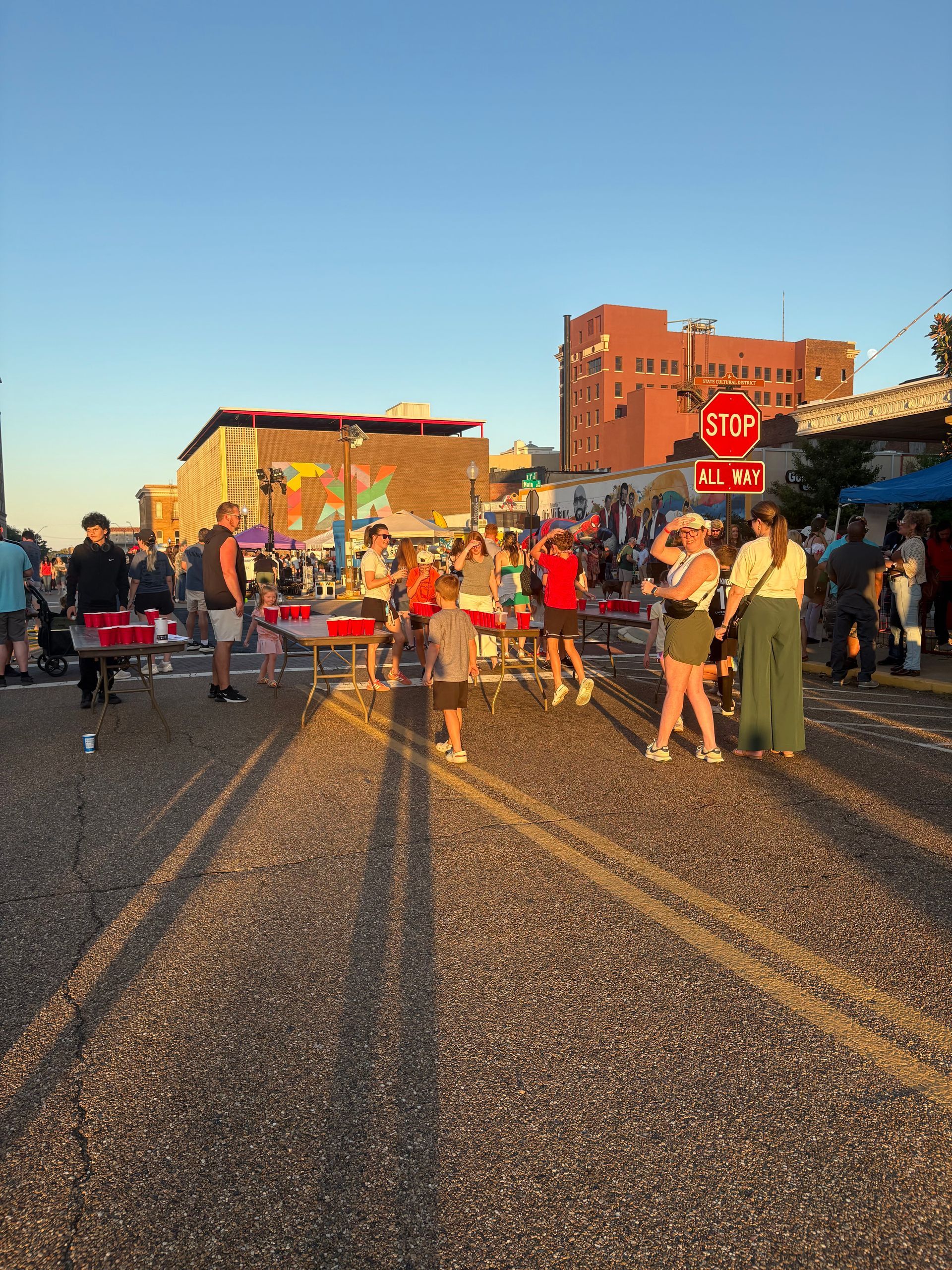 People gather outdoors with tables and buildings in the background during the sunset.