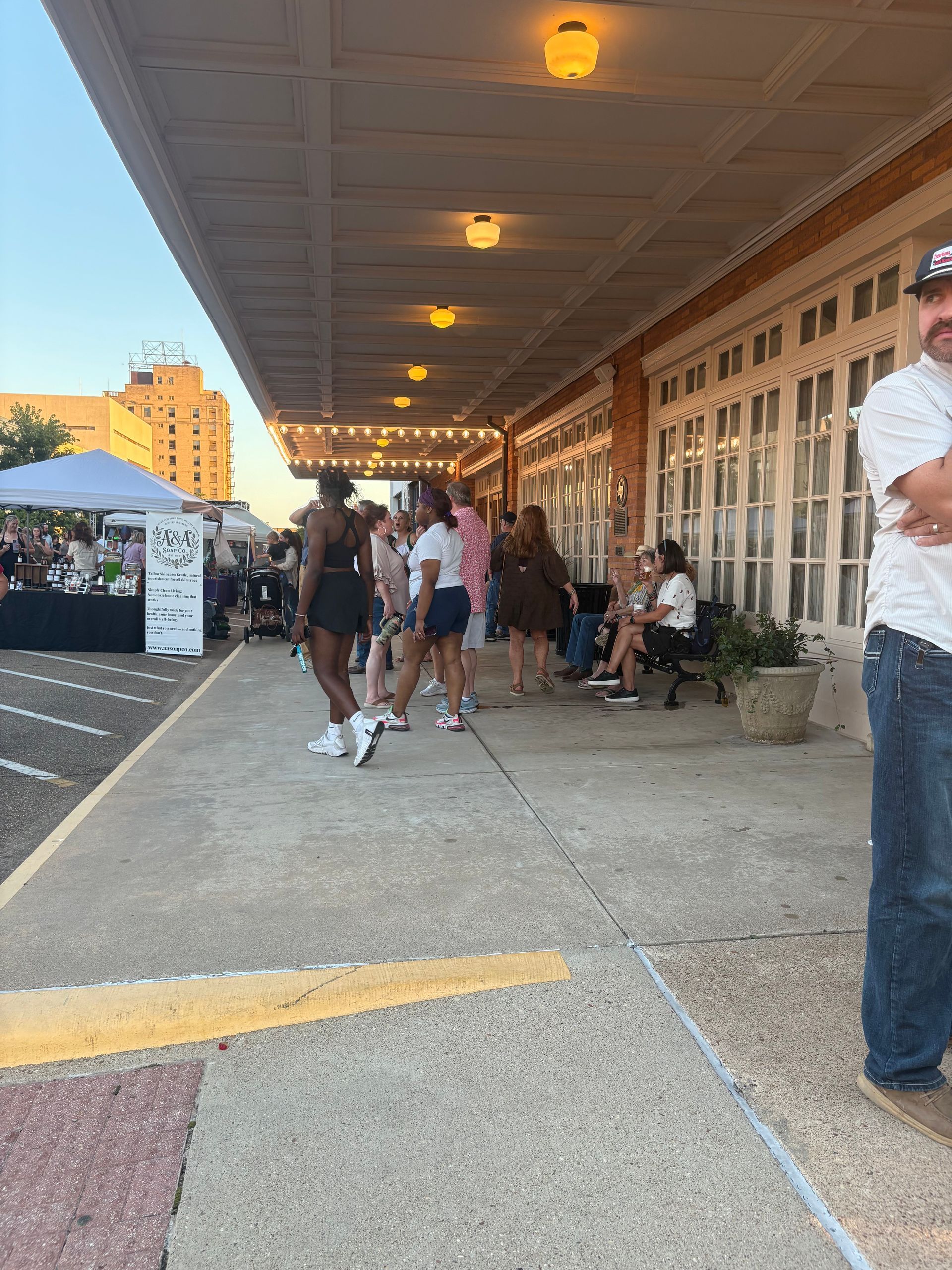 People gather under a building's awning. A few stand on a sidewalk, while others sit. Evening light.