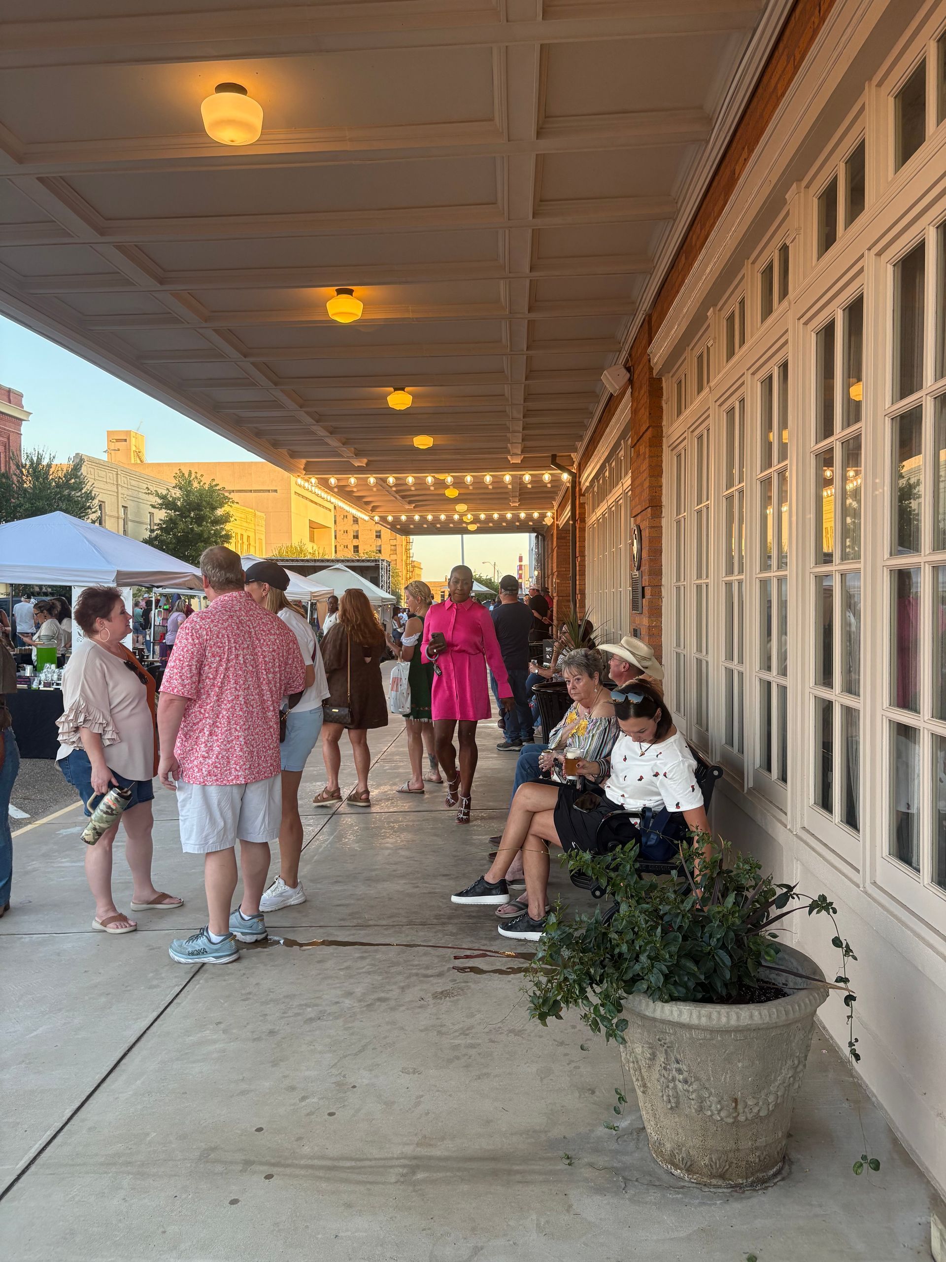People gather under a building's awning. Some stand, others sit, with potted plants. Evening lighting.
