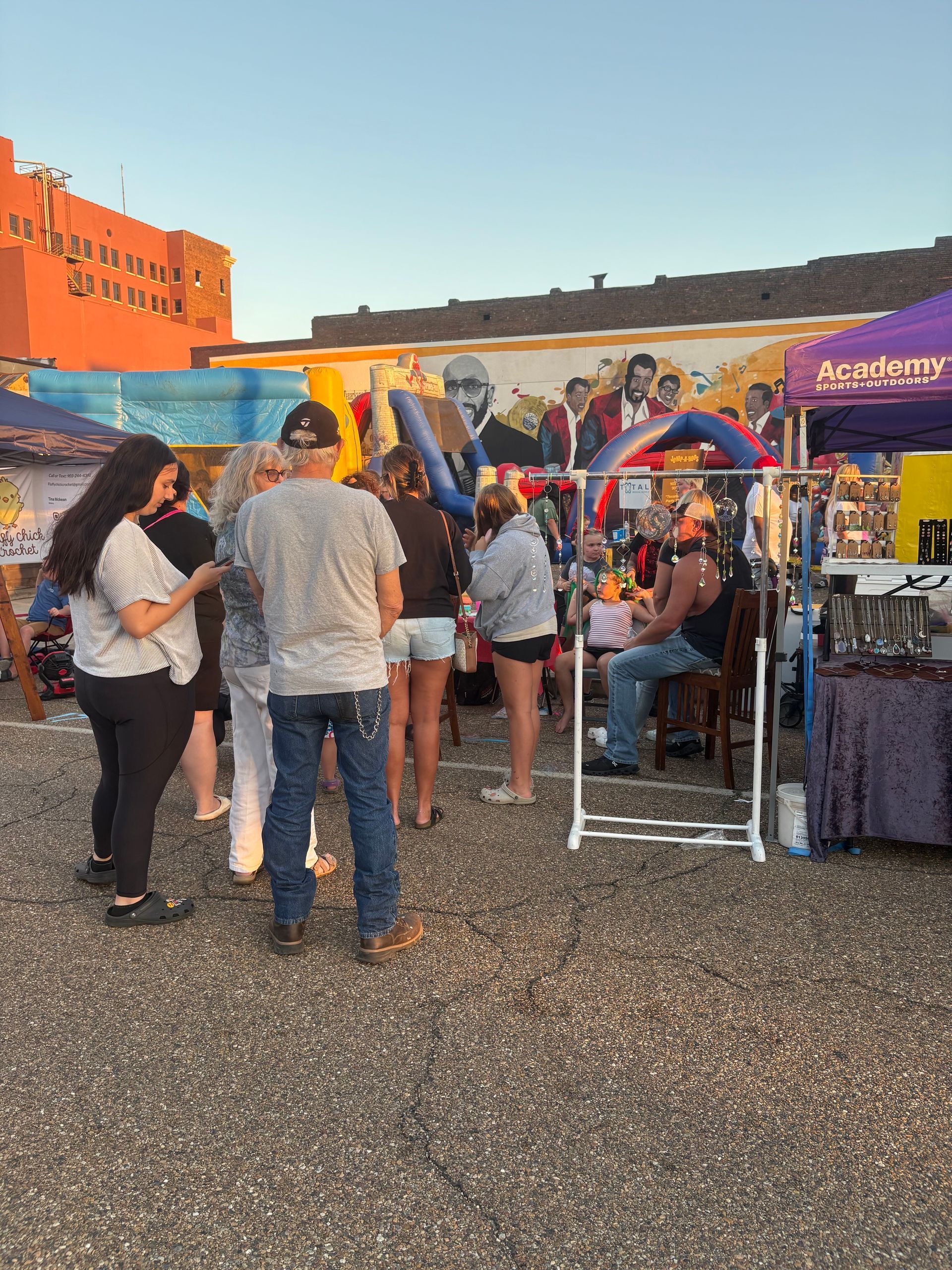 Outdoor event with vendor tents and people milling about, against a mural backdrop.