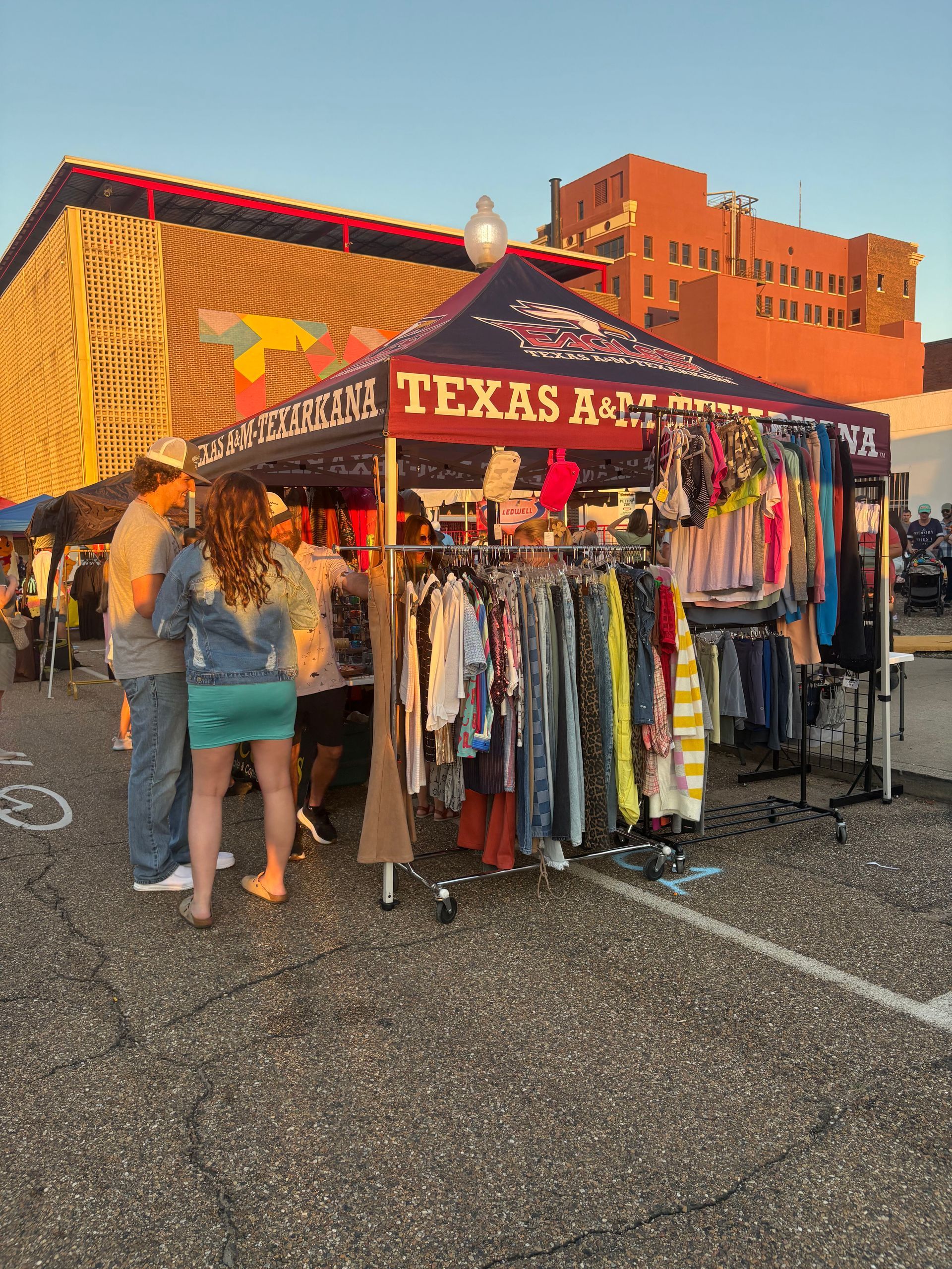 A clothing stall under a Texas A&M tent, with people browsing items, set against a city backdrop.