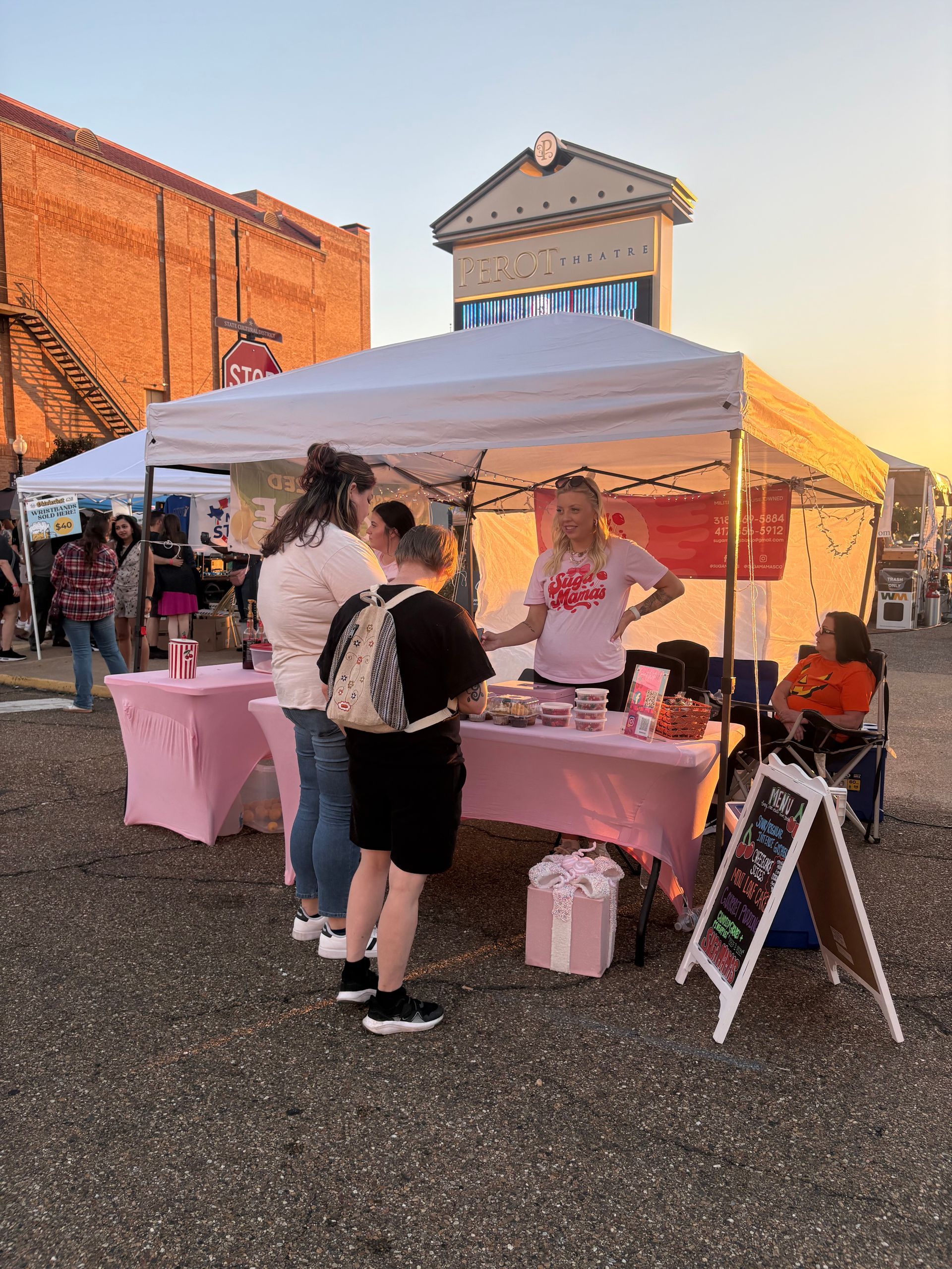 Vendor at outdoor event selling items at pink table under white tent; customers browsing.