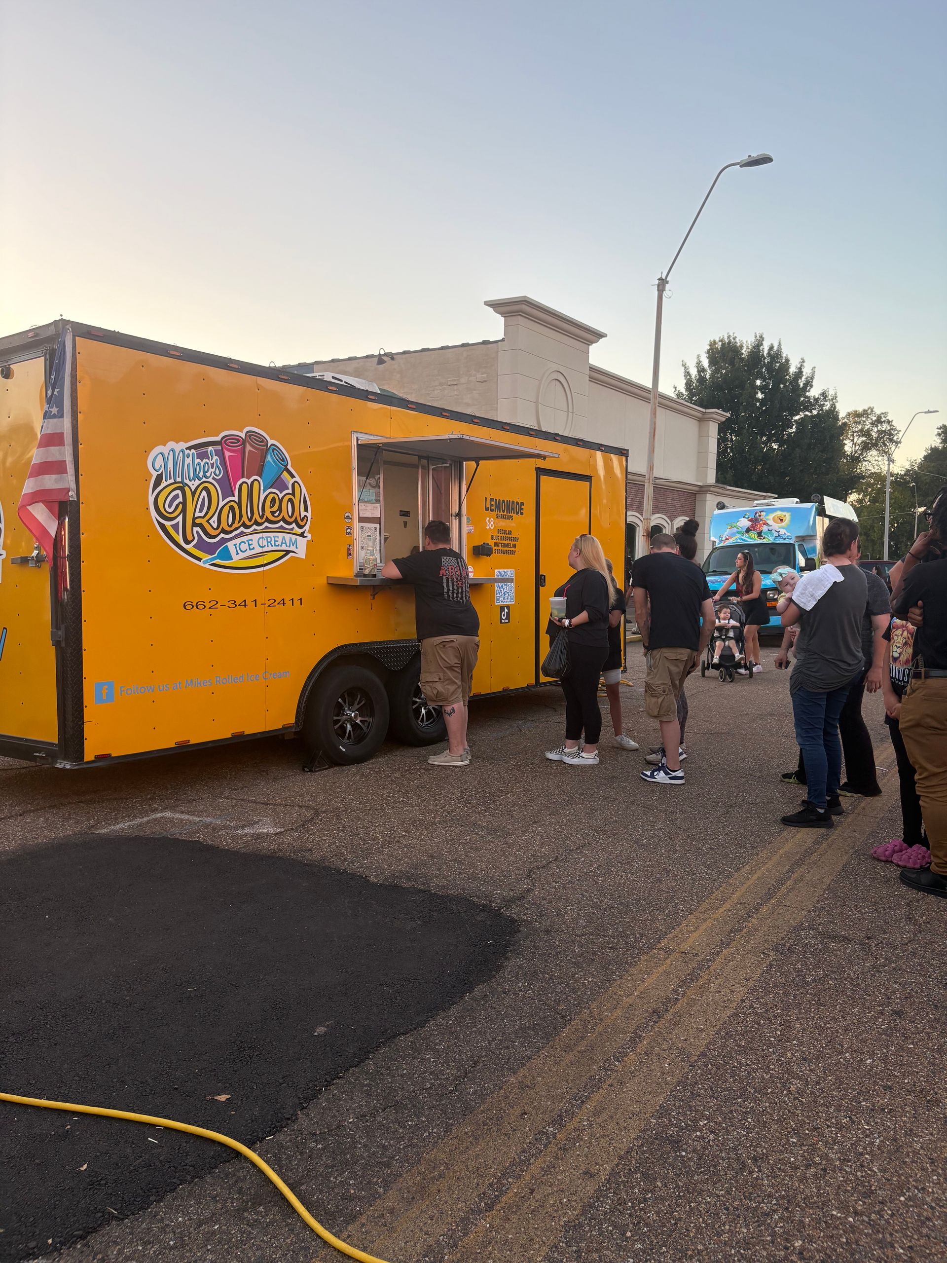 Yellow food truck with a line of people ordering. An American flag hangs on the side. Evening setting.