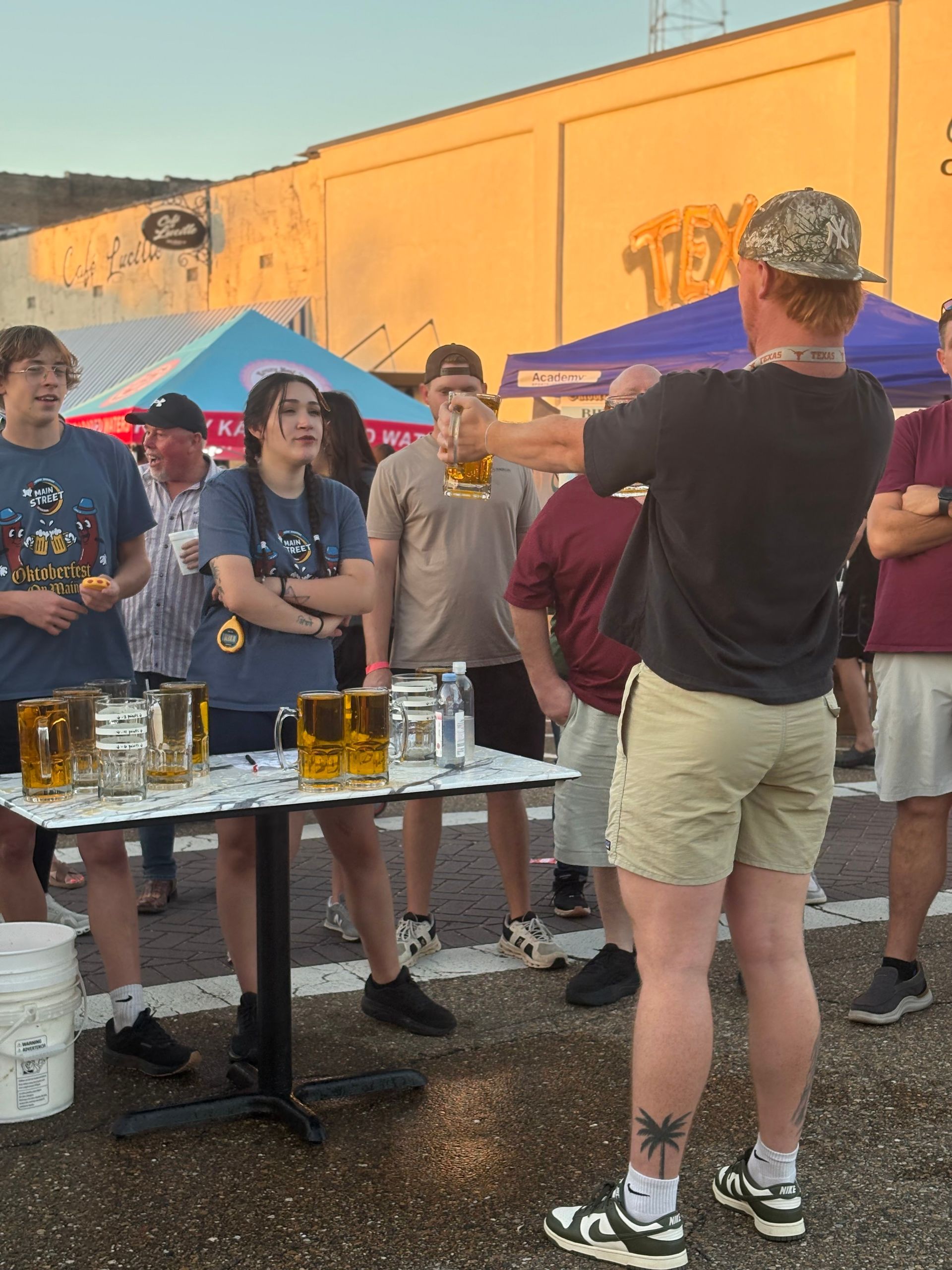 People at an outdoor beer-holding contest, some with arms crossed, others watching. Setting is a street.