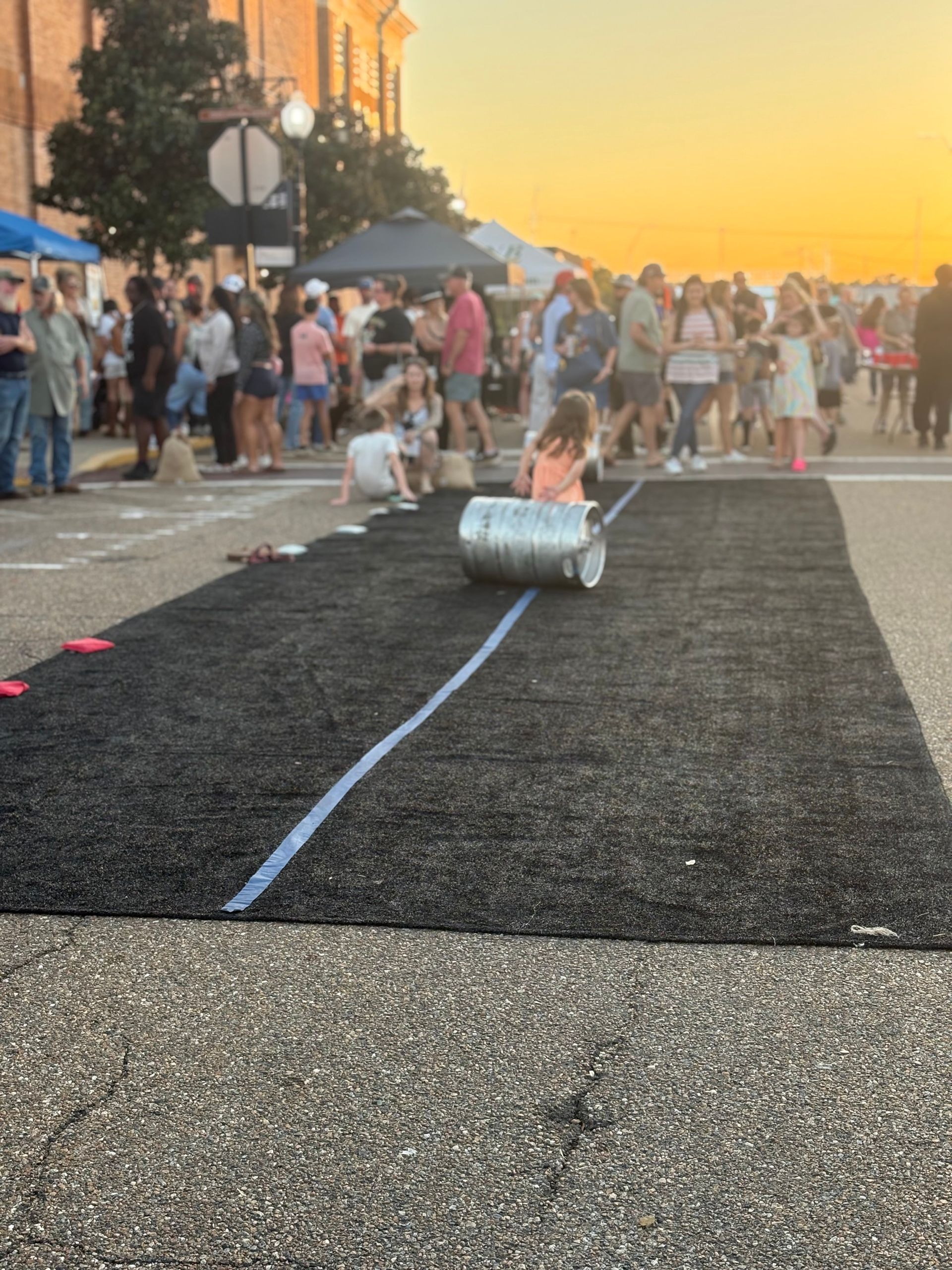 People watch children rolling a metal barrel down a black track on a street at dusk.