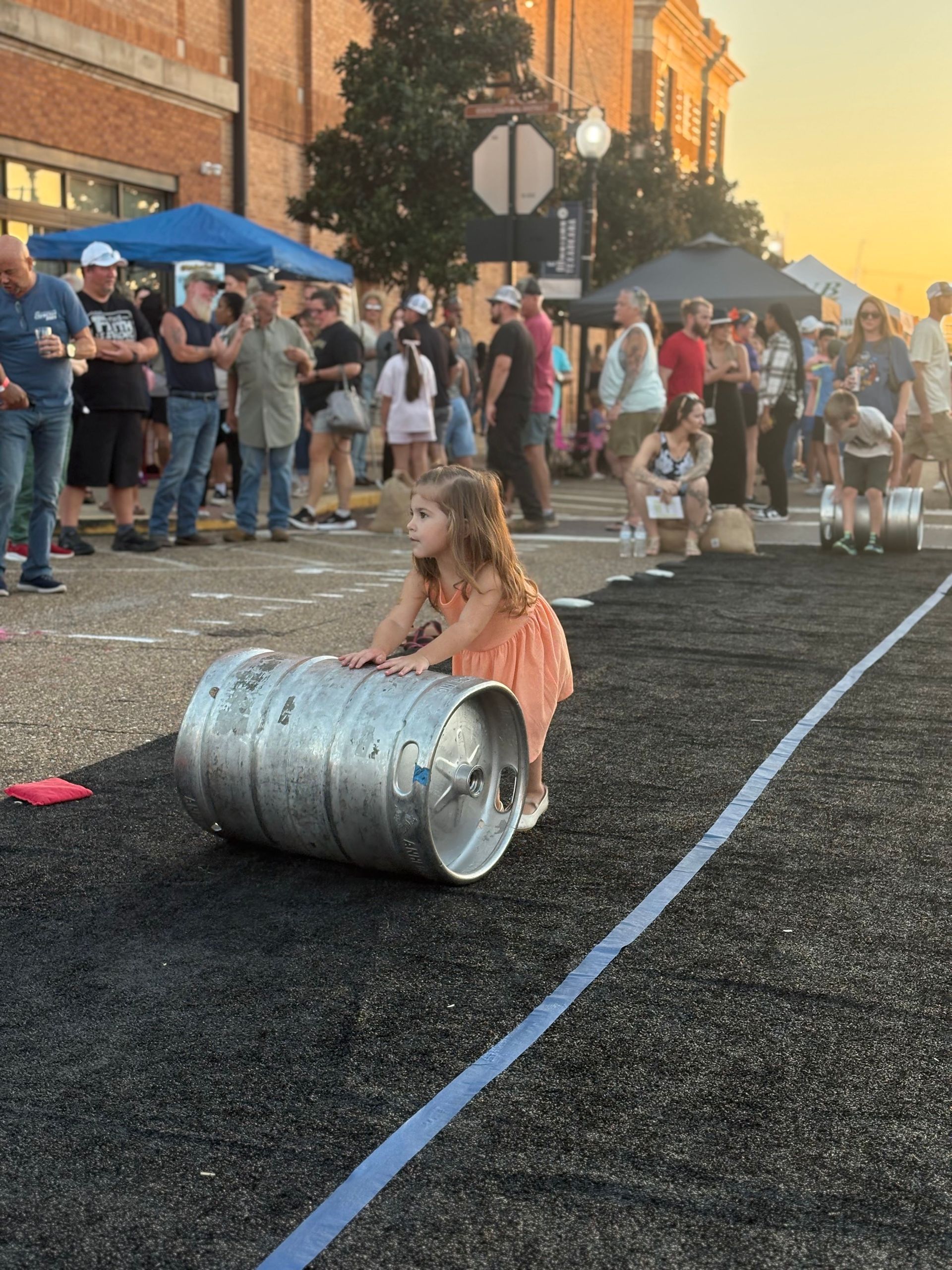 Girl rolling a beer keg on a track during an outdoor festival, crowd in the background.