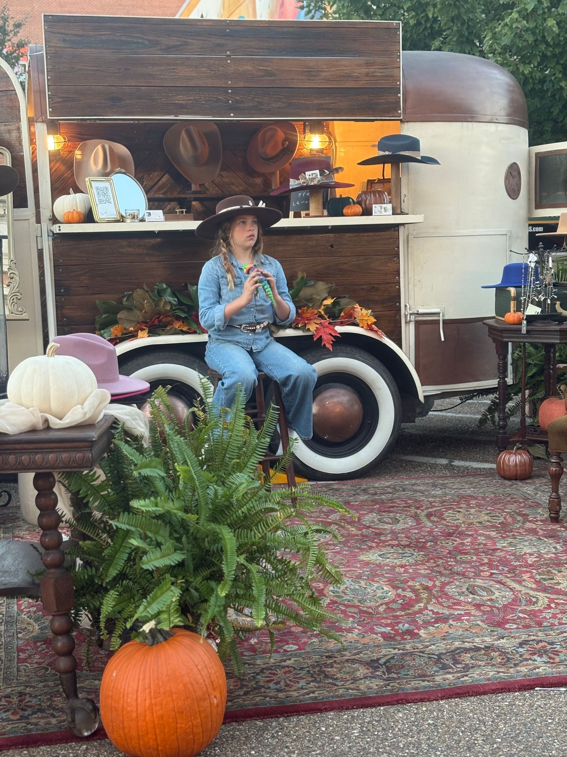 Person in denim jumpsuit sits near a trailer decorated for autumn. Pumpkins, hats, and plants are visible.