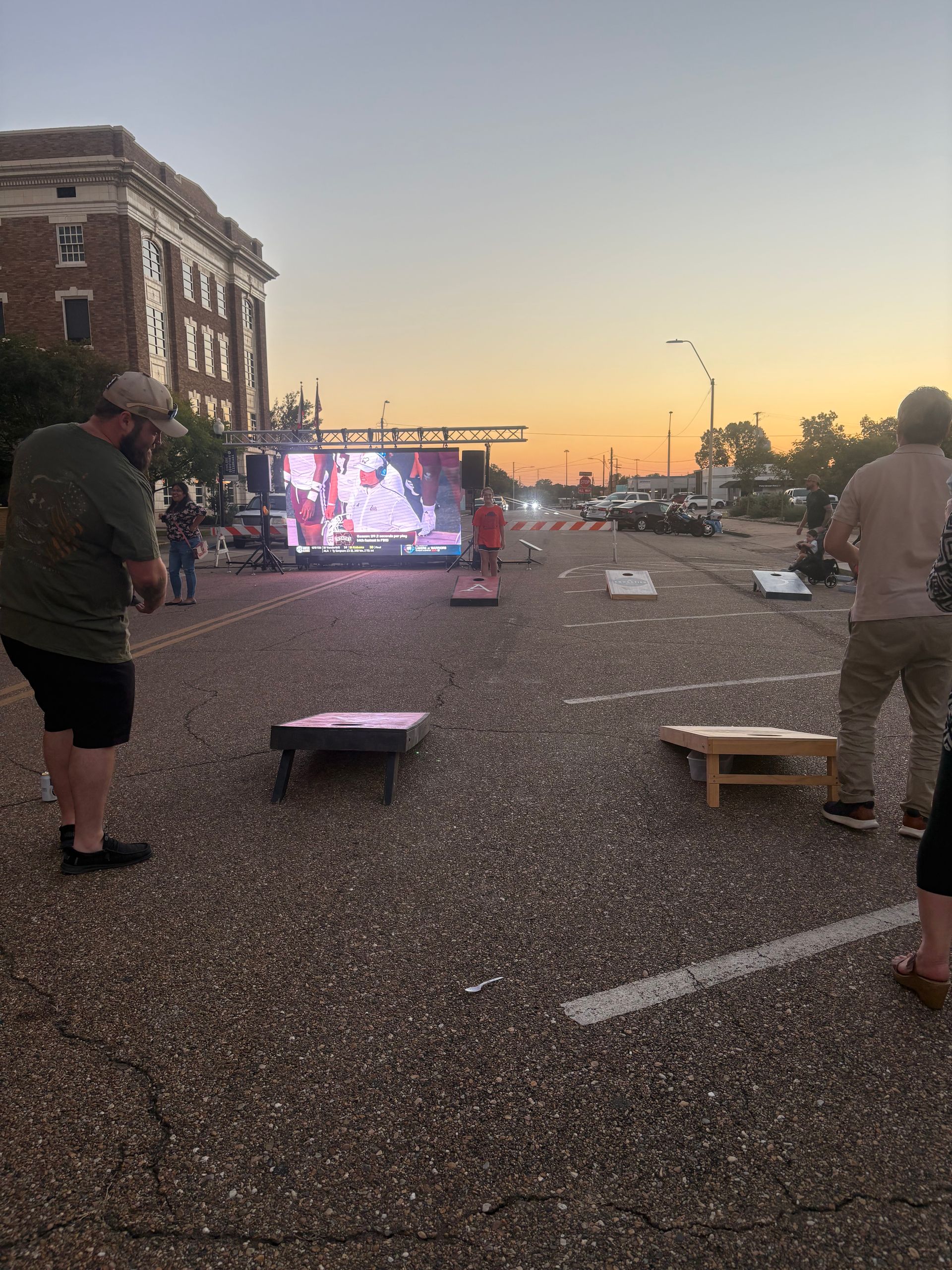 People playing cornhole in a street at dusk. A large screen displays an image.