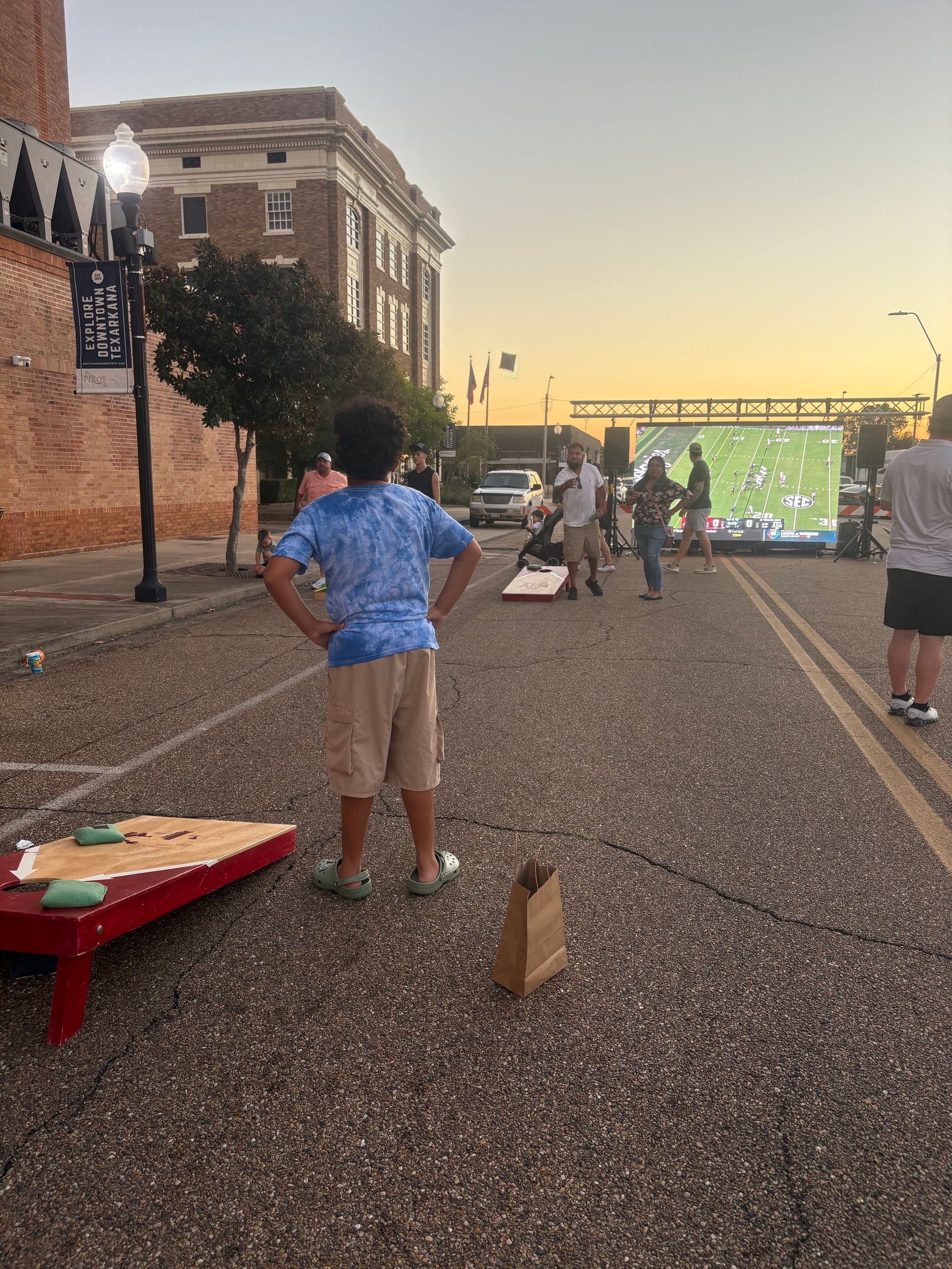 Boy in blue shirt watches outdoor movie and plays cornhole. Sunset in background.
