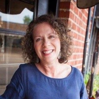 Woman with curly brown hair smiles, wearing a blue top, standing outdoors near brick.