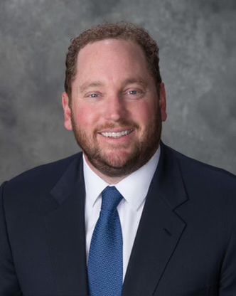 Smiling man in a dark suit, white shirt, and blue tie against a gray studio backdrop