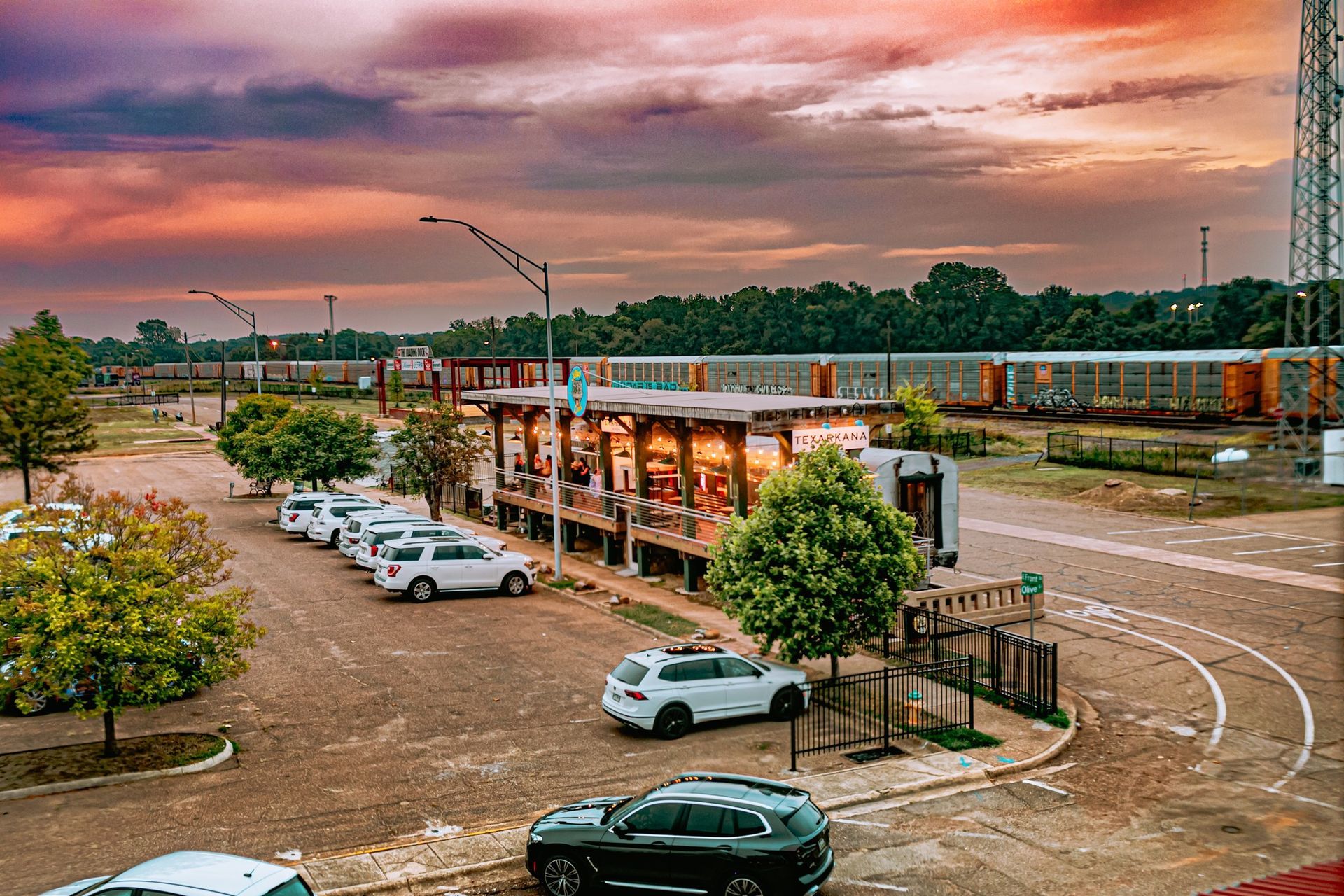 Outdoor venue with cars parked, trees, a building, and a sunset sky.