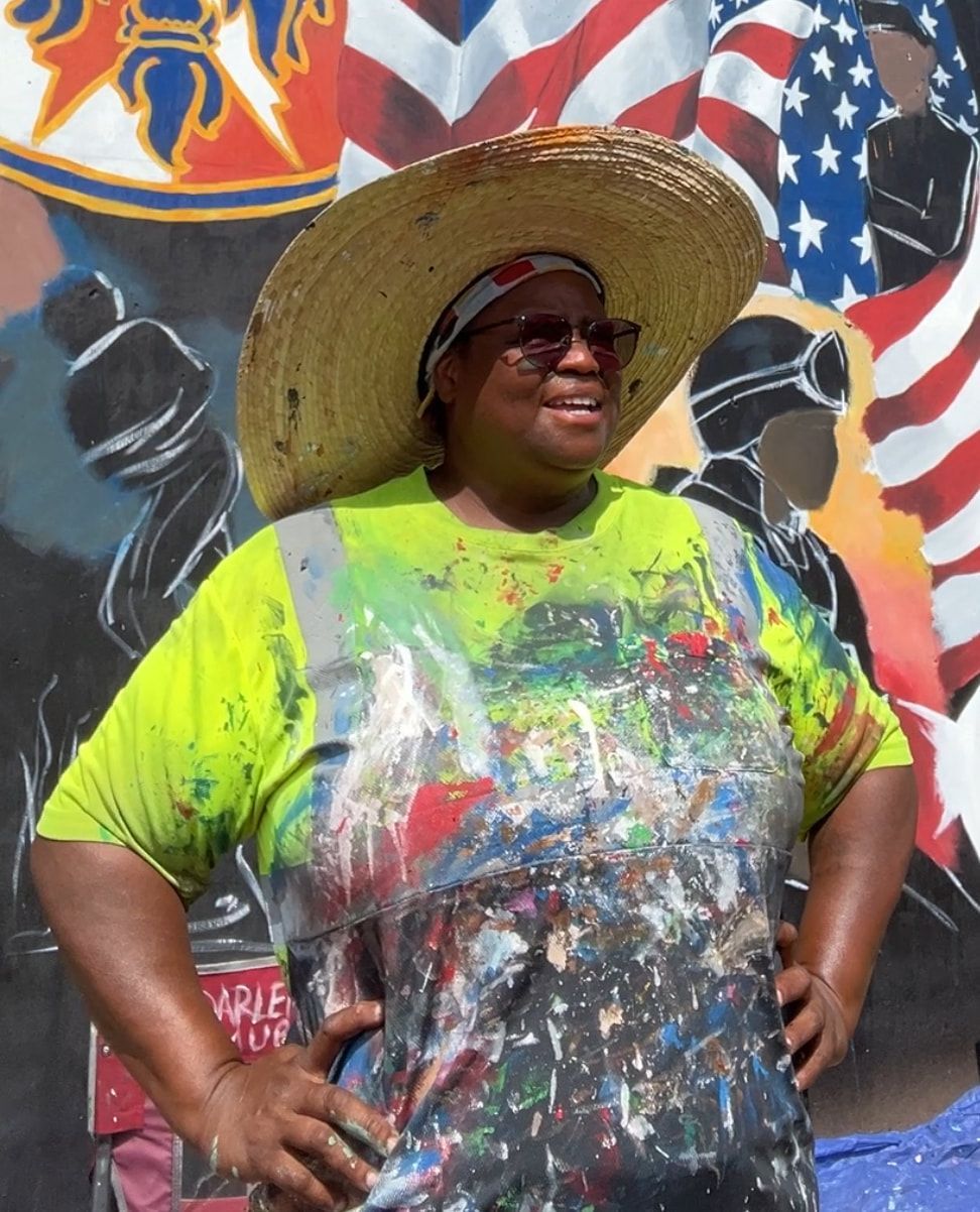 Woman in paint-splattered shirt and straw hat poses in front of a colorful mural, with an American flag visible.
